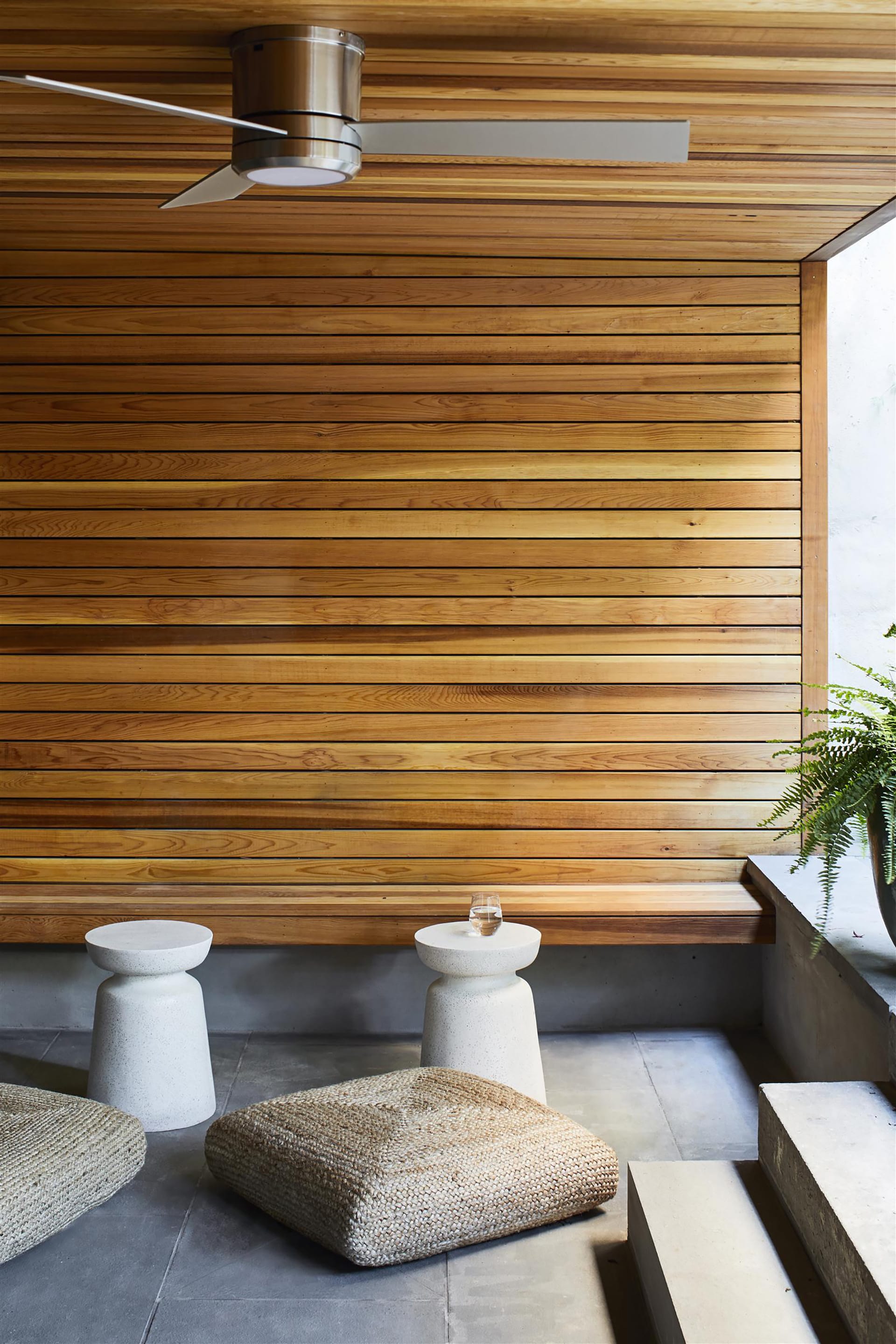 Areaway leading to a rear yard with two white side tables, wood paneled walls and ceiling, a modern ceiling fan, bluestone pavers, and two cushions.