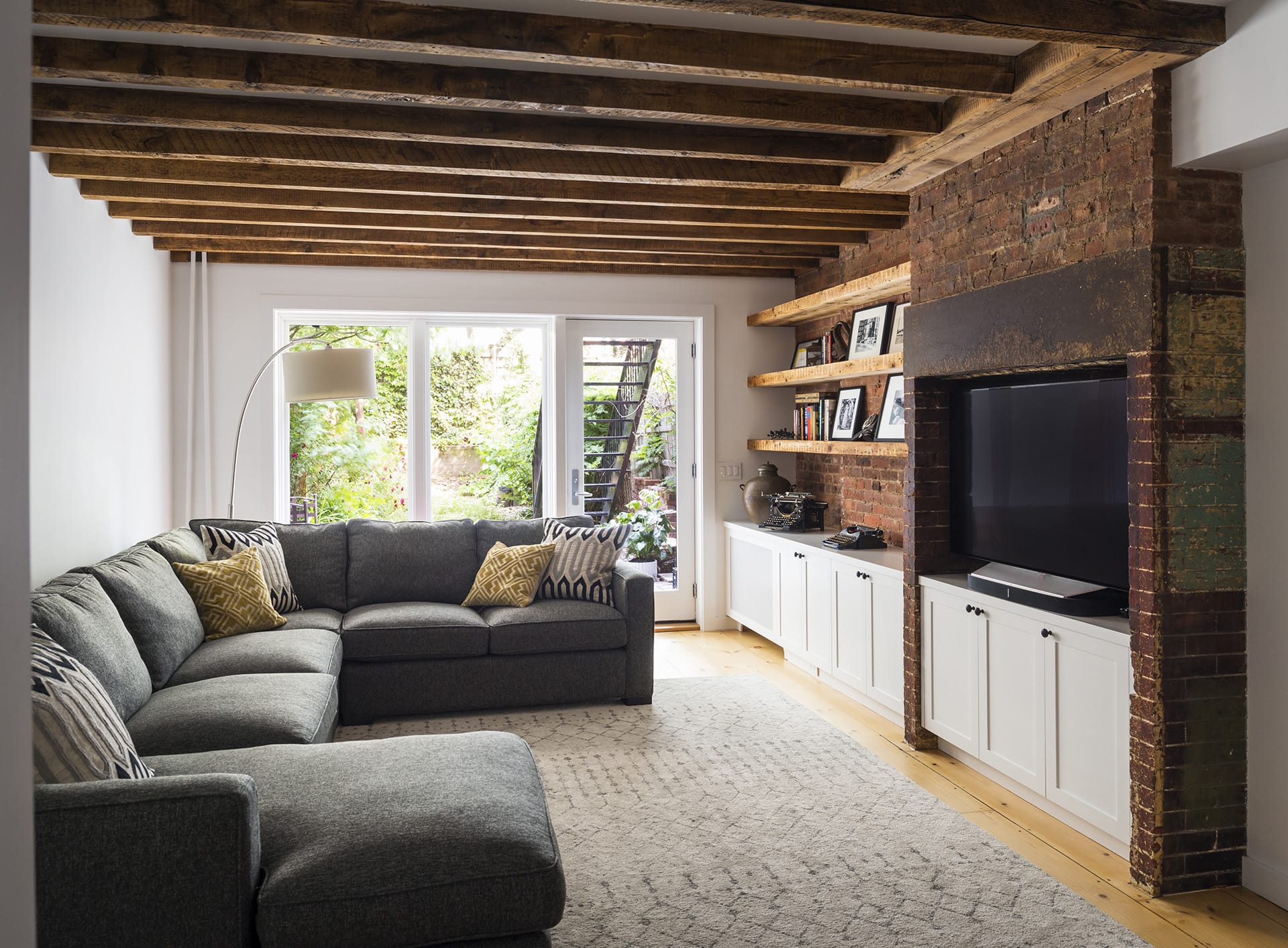 Renovated garden floor of a Cobble Hill townhouse with large glass doors, exposed joists, shelves, couch and tv.