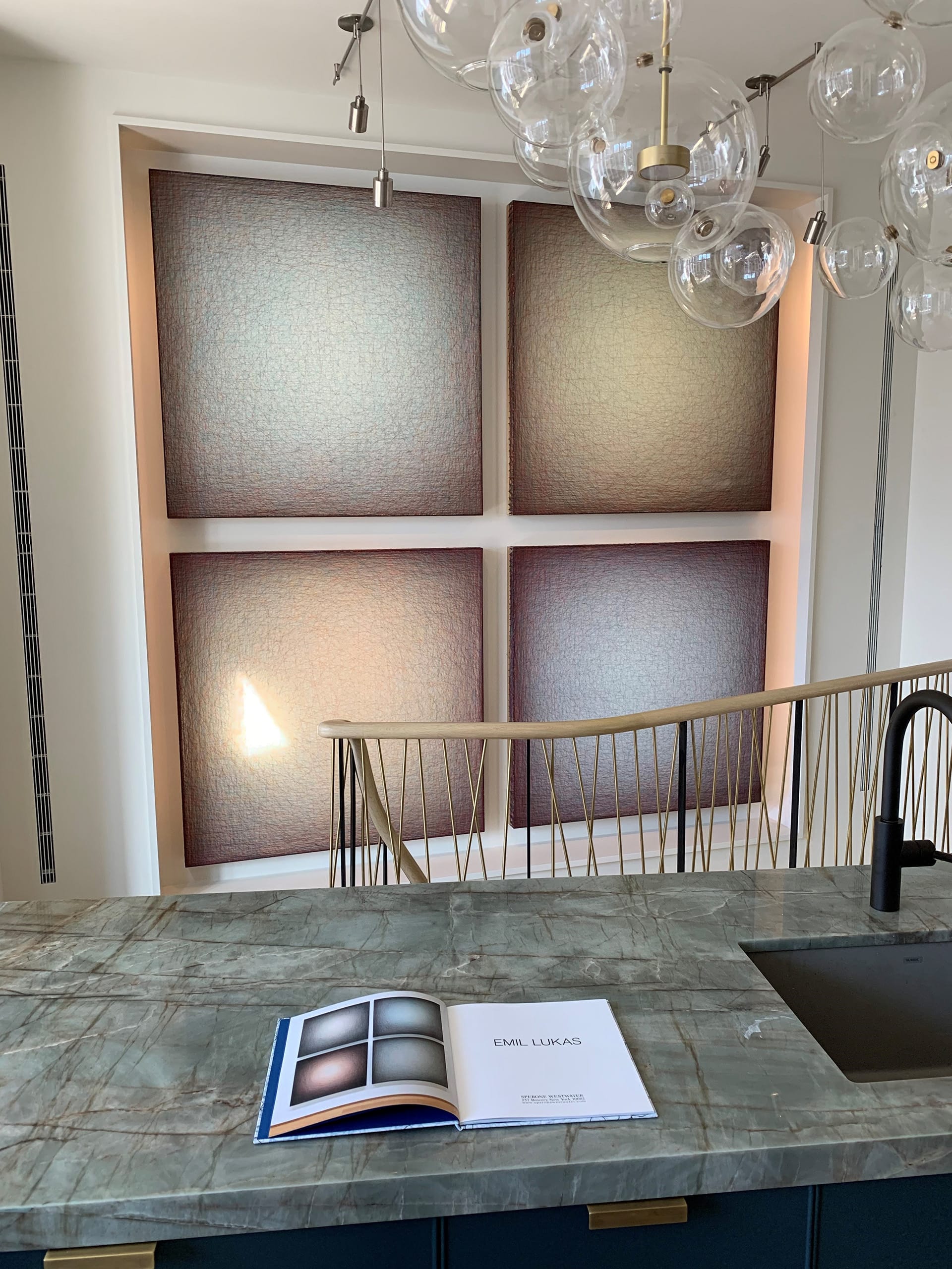 Four-panel multimedia artwork hanging on a wall above a staircase. A glass globe chandelier and a green stone counter are in the foreground.