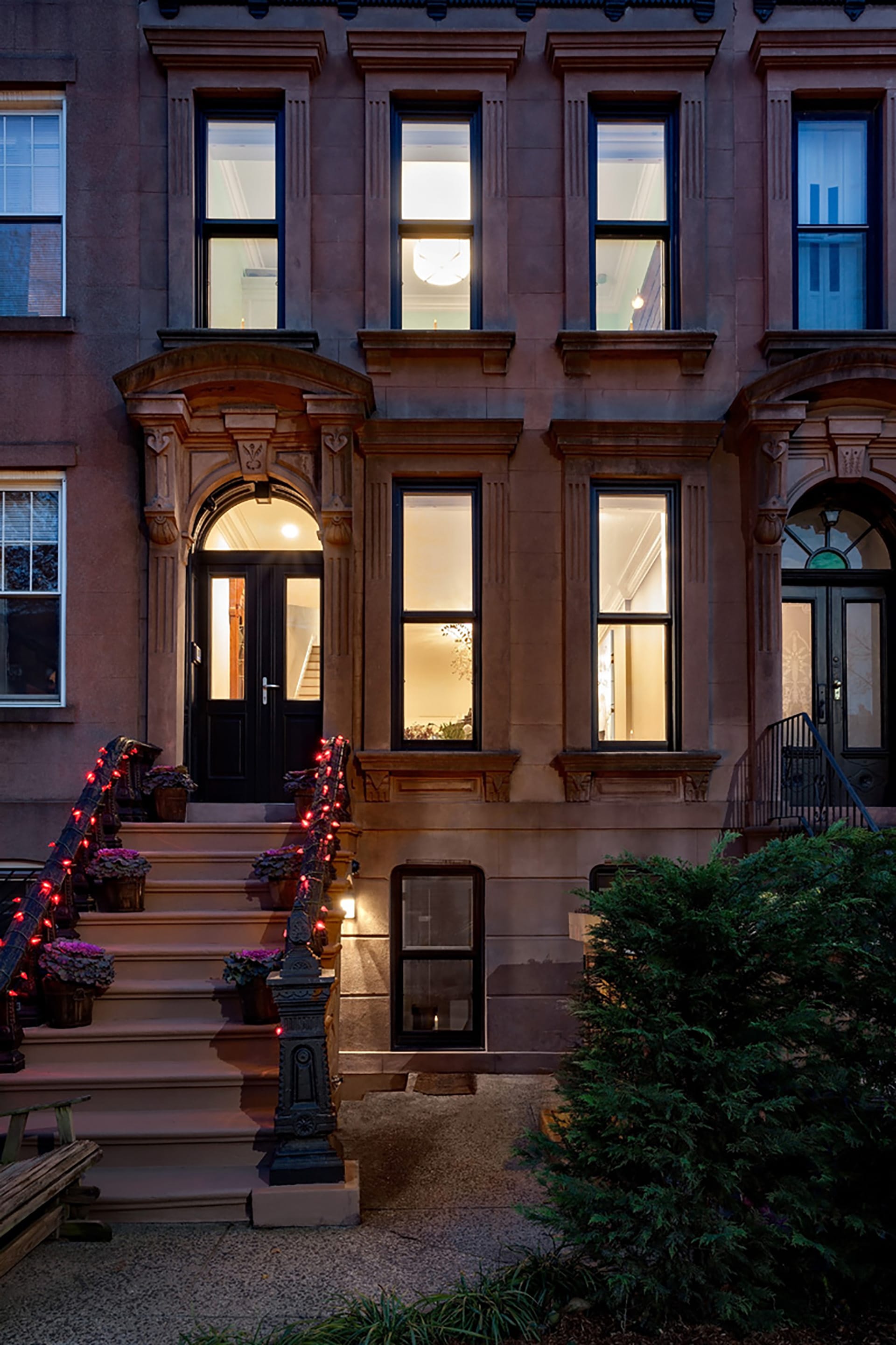 Front façade of a Carroll Gardens brownstone with red Christmas lights lining the stoop, at twilight.