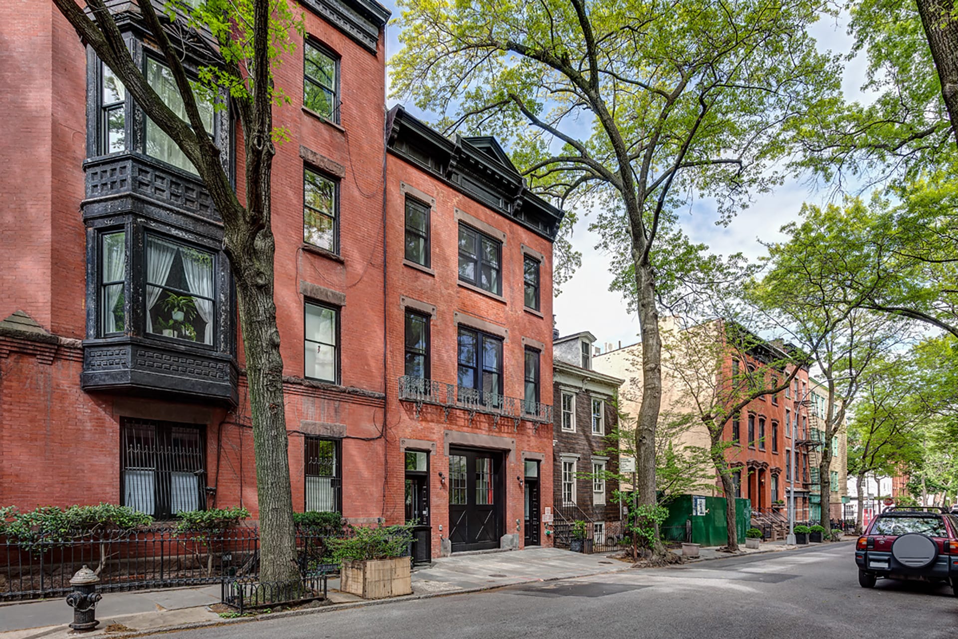 Front facade of a three-story brick rowhouse with black doors, a black cornice, and brownstone accents. A large tree sits on the sidewalk in front of it.