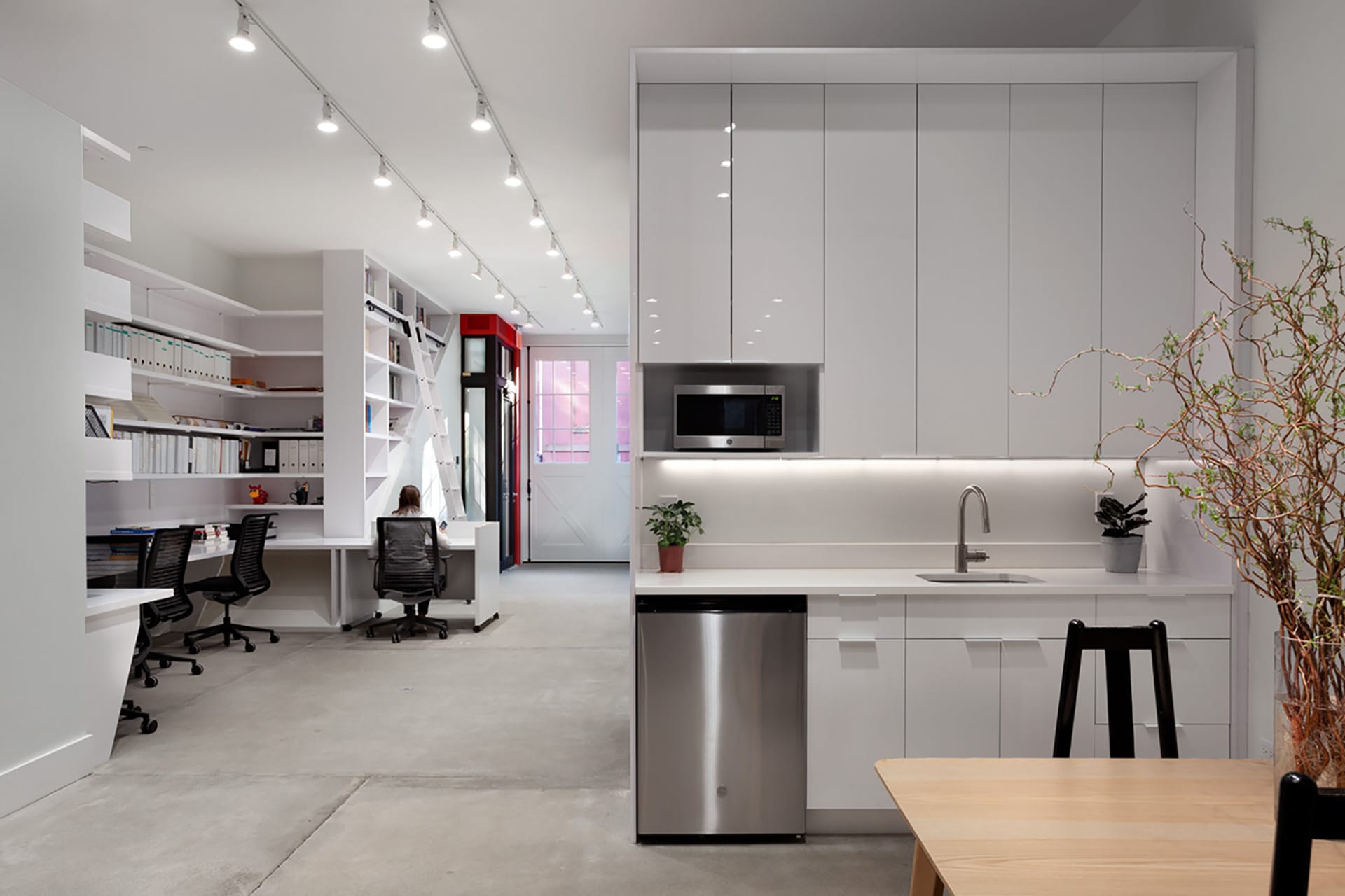 All-white kitchenette and office space with cement floors and a pop of red paint surrounding the front door.