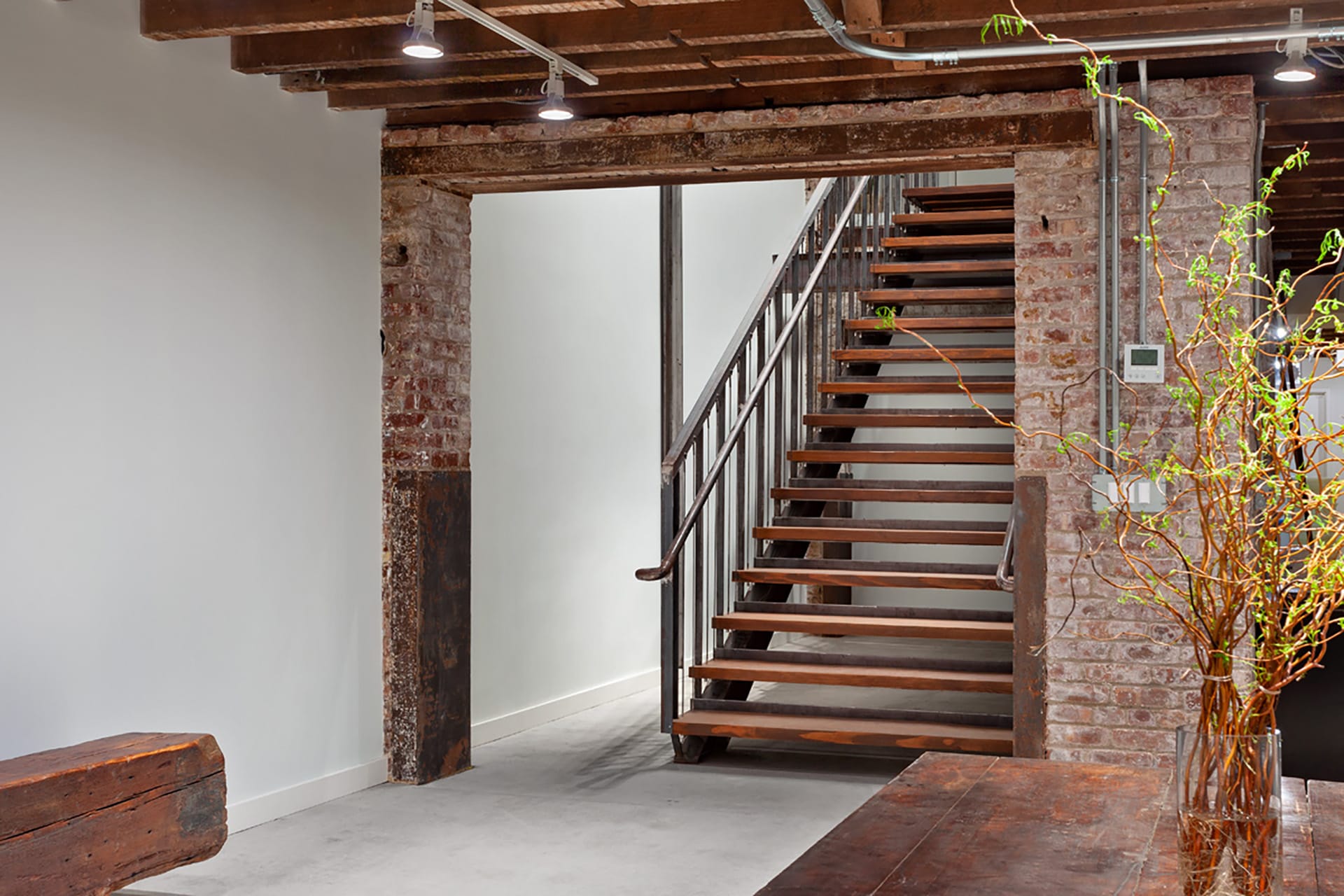 Staircase leading to a cellar with exposed ceiling beams, antique bench, and antique table with a vase and plants on top of it