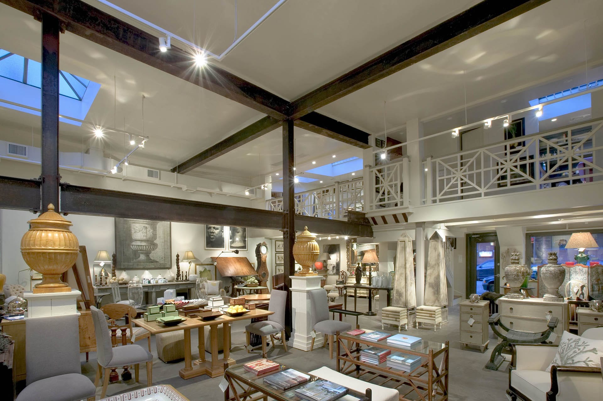 Photo of the inside of an antique shop. The walls and ceilings are painted white, large aged metal beams are both decorative and supportive, and a mezzanine with spiderweb-like railings is visible at the top right of the photo.