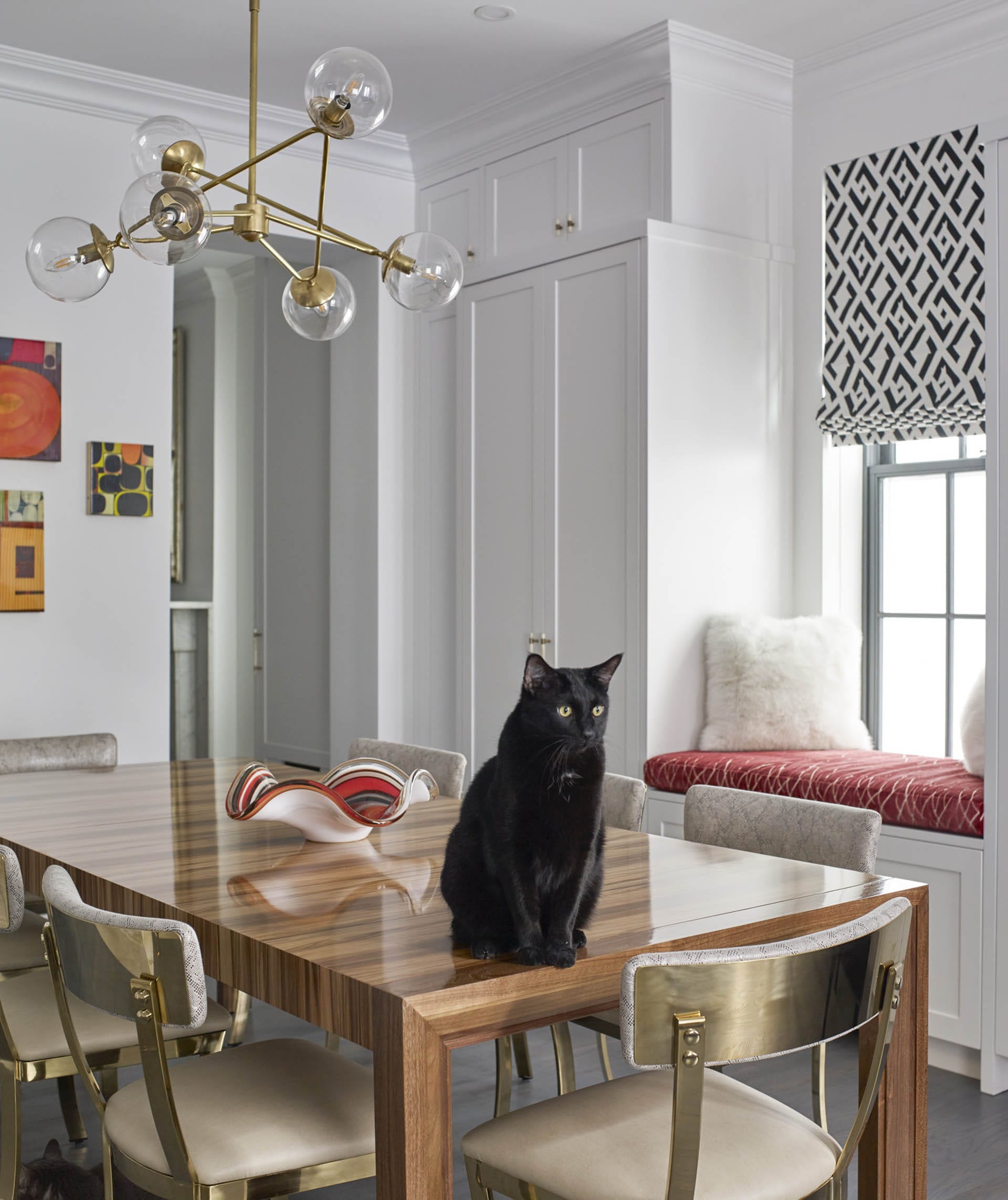 Dining room with a wood table, klismos chairs, a gold chandelier, and a black cat perched on the table.