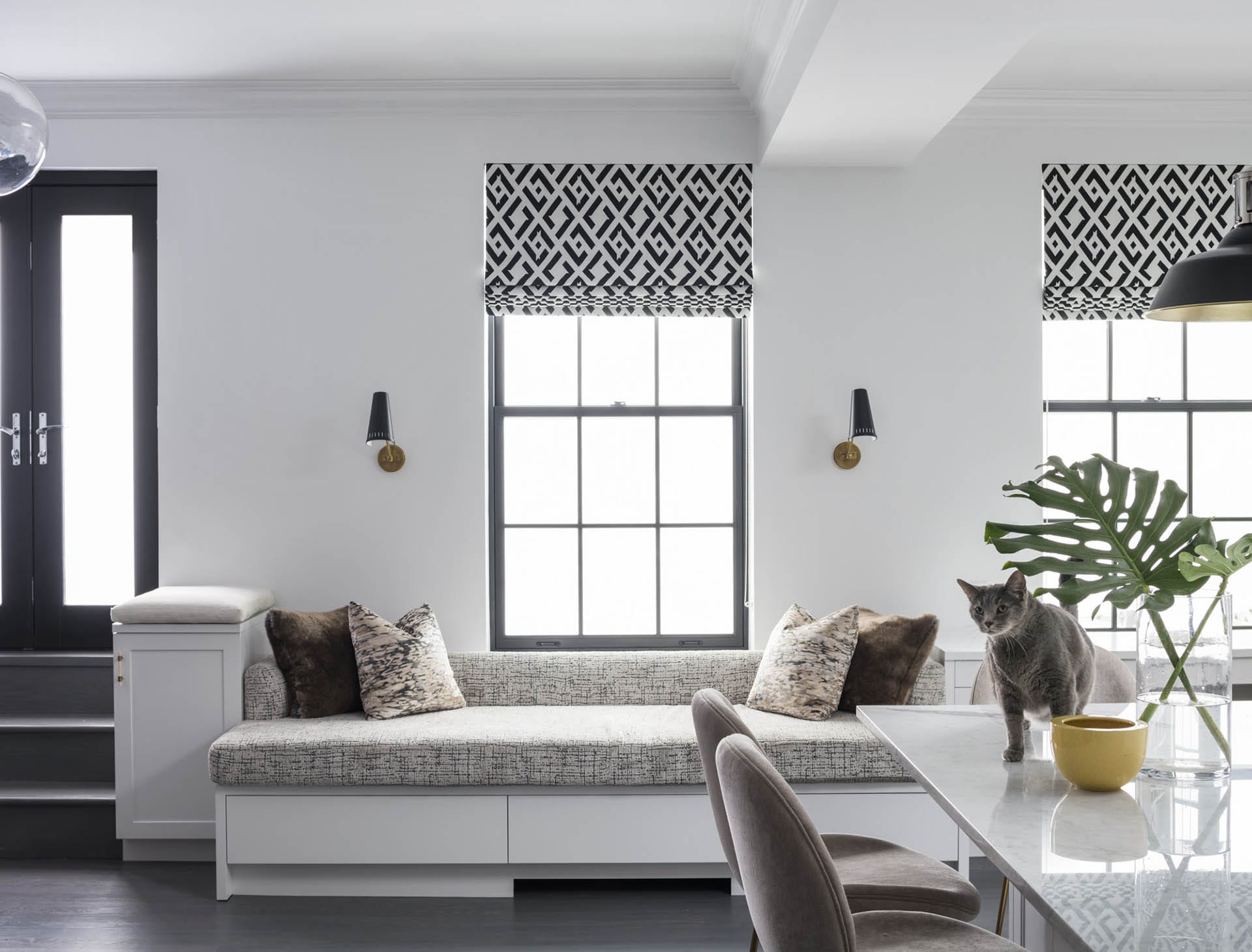 Living room with black French doors leading to the deck, black and white Roman shades, and a built-in bench. A grey cat stands on the kitchen table.