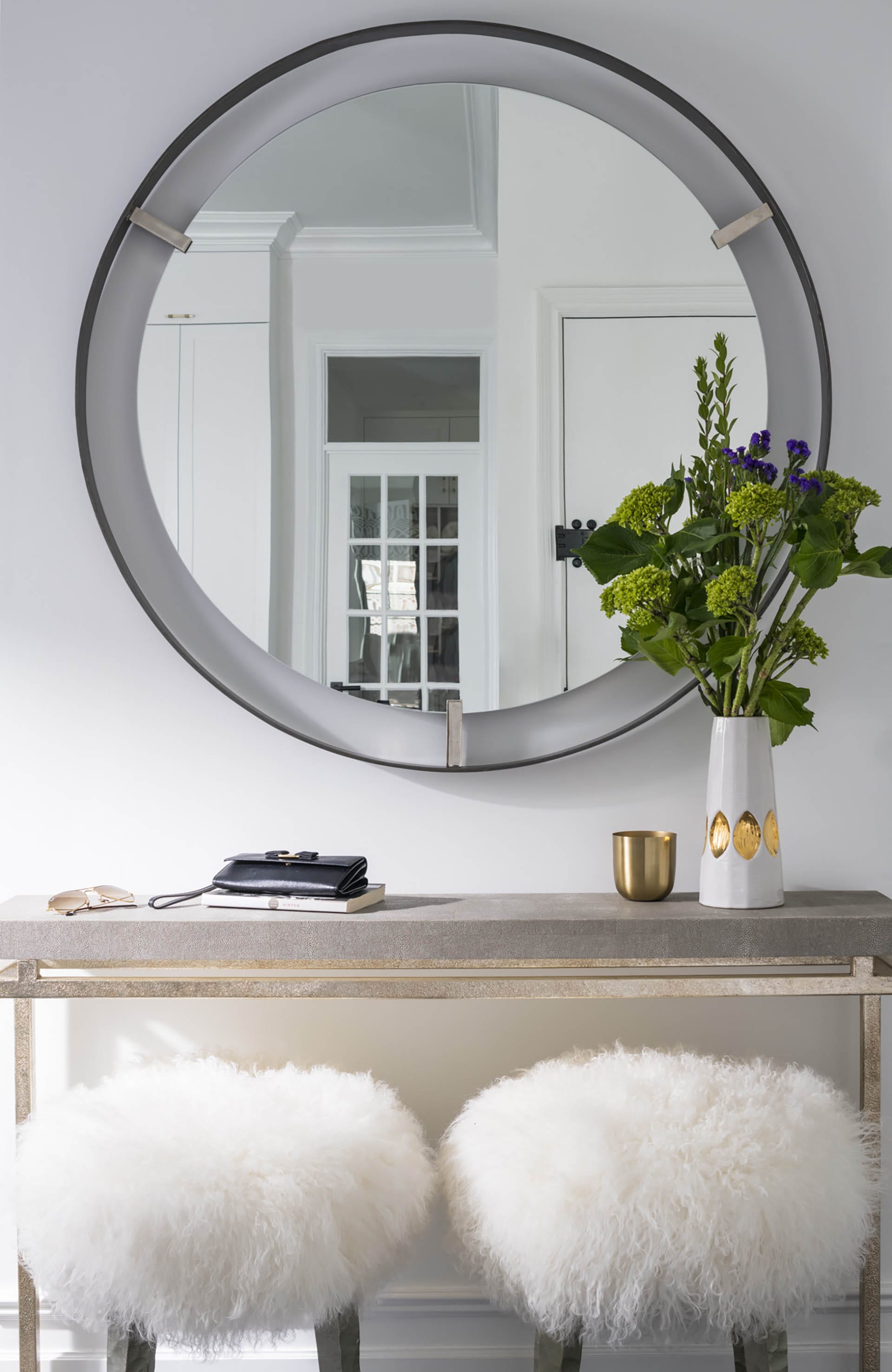 A circular mirror hangs above a credenza with two white, fuzzy stools in front of it.