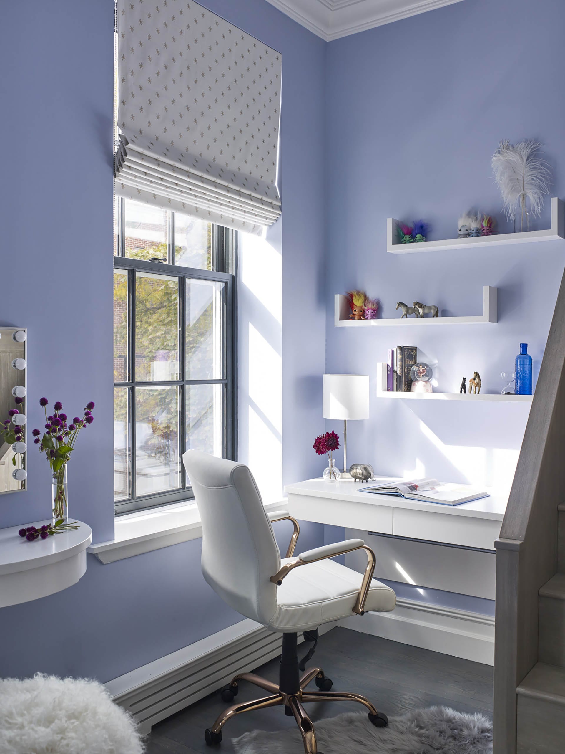 Office with lavender walls, white desk and floating shelves, and white Roman shades