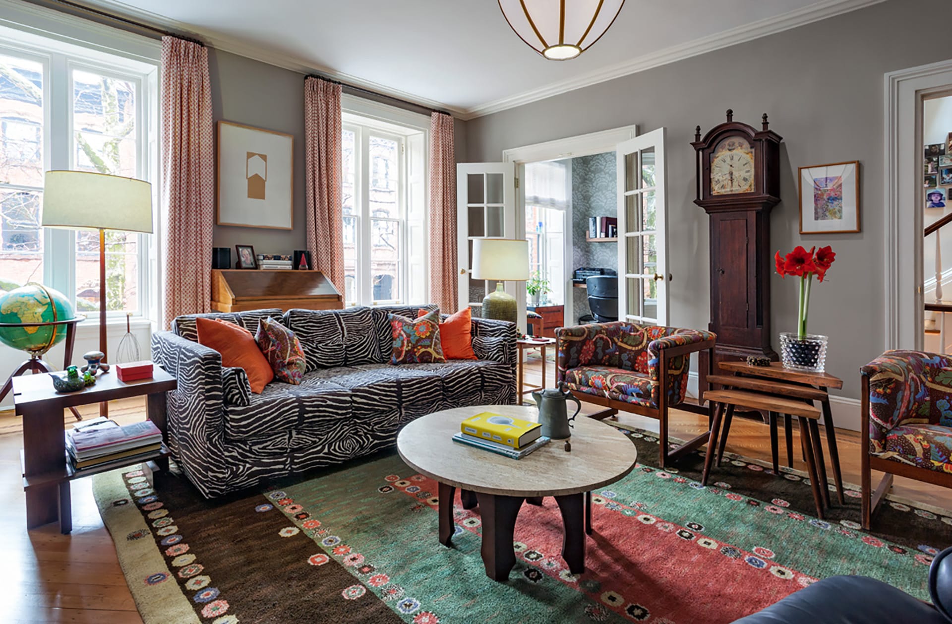 Bright, eclectic living room with patterned couches and chairs, a large patterned rug, light grey walls, and a Grandfather clock.