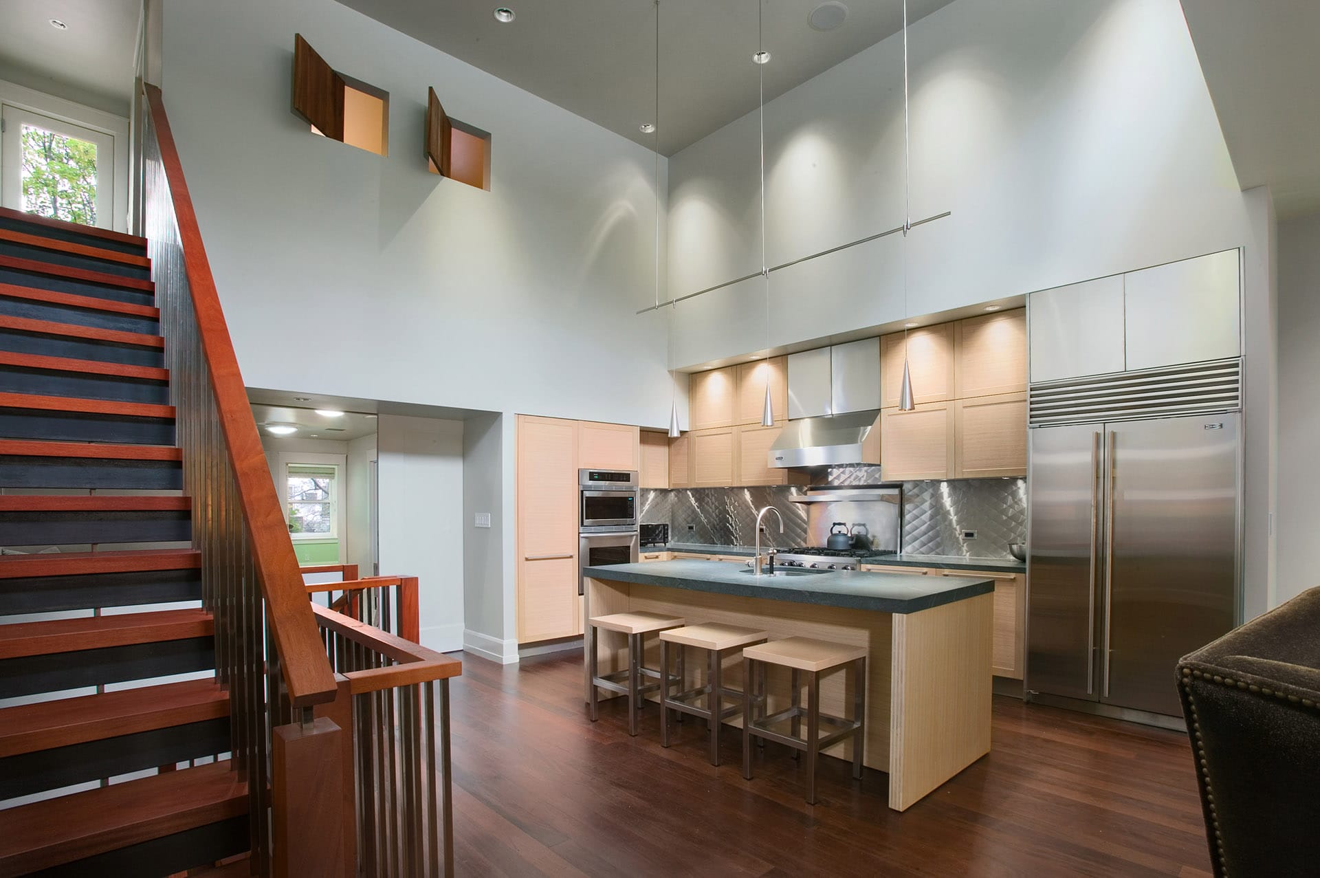 Modern double-height kitchen with dark wood floors, light wood cabinetry and island, and stainless steel backsplash and appliances.