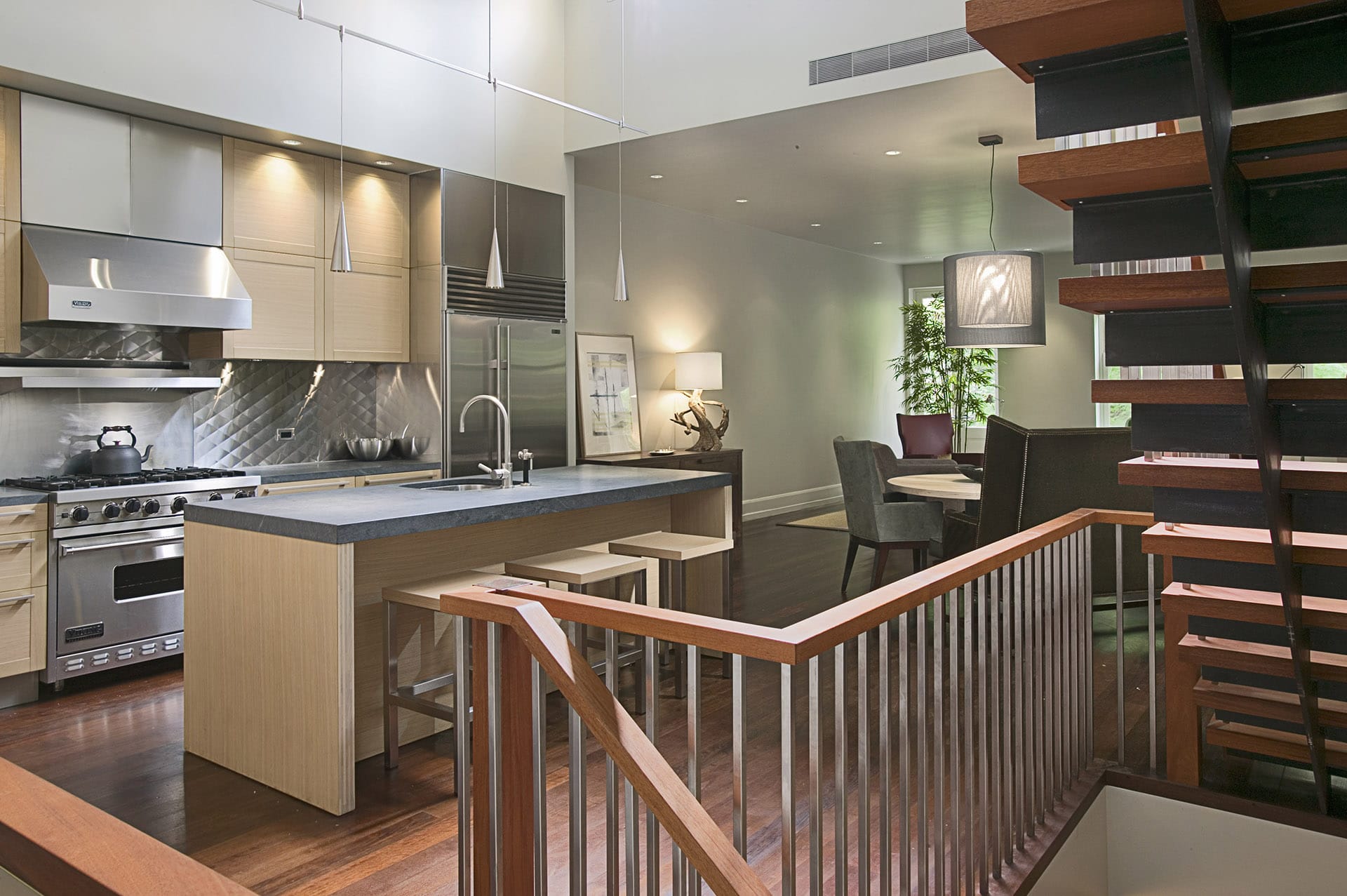 Modern double-height kitchen with dark wood floors, light wood cabinetry and island, and stainless steel backsplash and appliances. A open-stringer, open-riser modern staircase is at the right foreground.