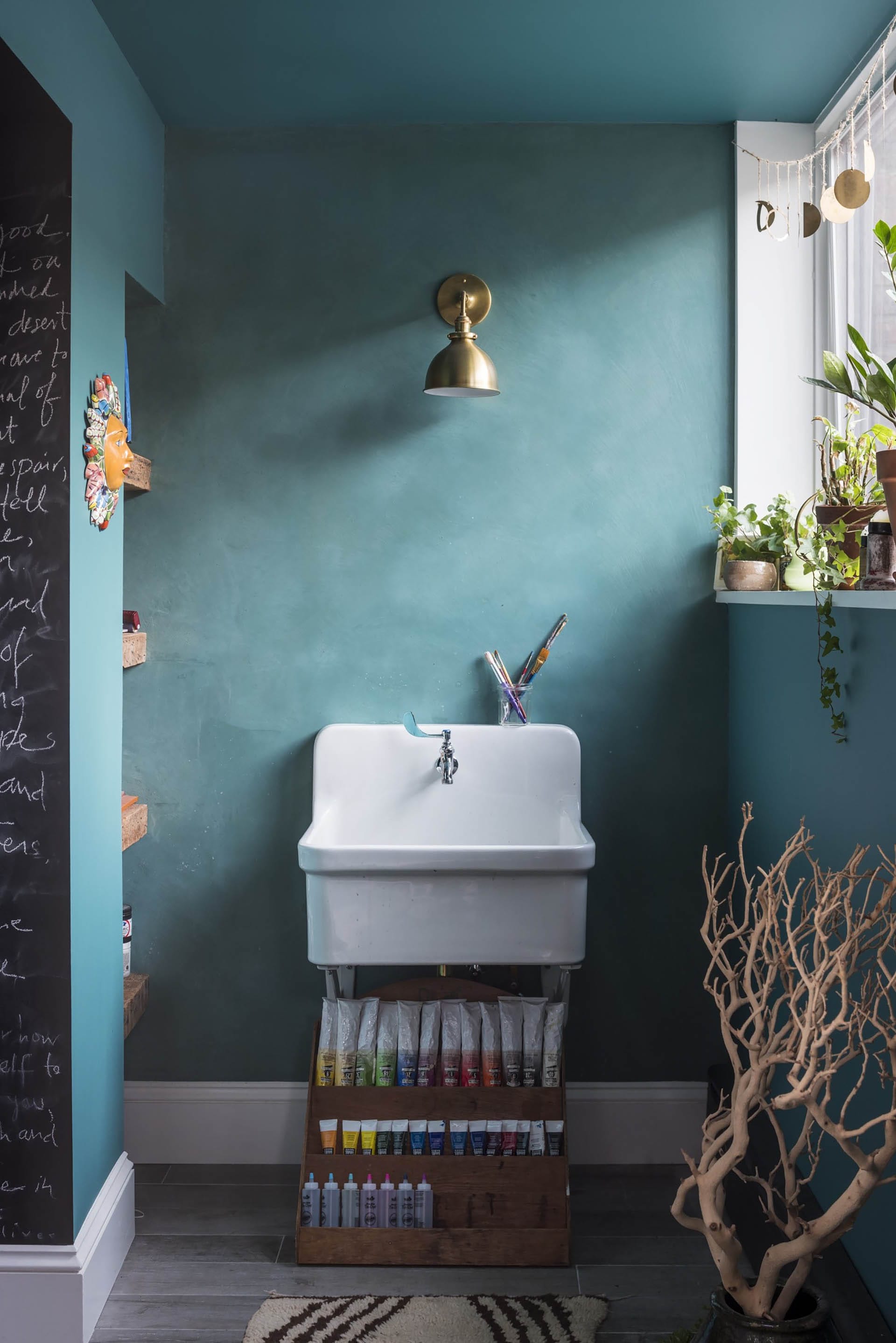 Teal room with a gold sconce above a white sink and painting station. Several potted plants sit on the windowsill to the right.