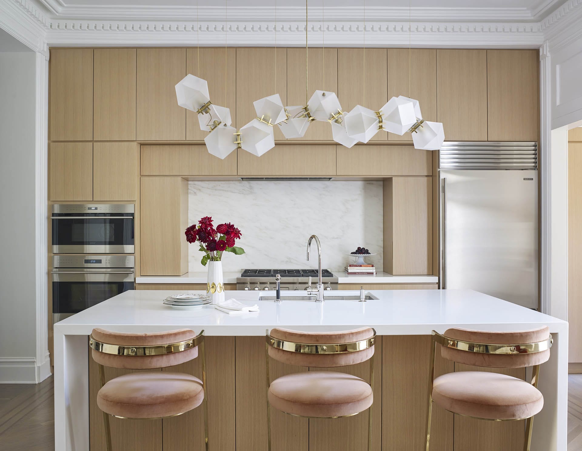 Kitchen with natural wood cabinetry, a white island and marble backsplash, restored historic crown molding, and three pink barstools at the island.