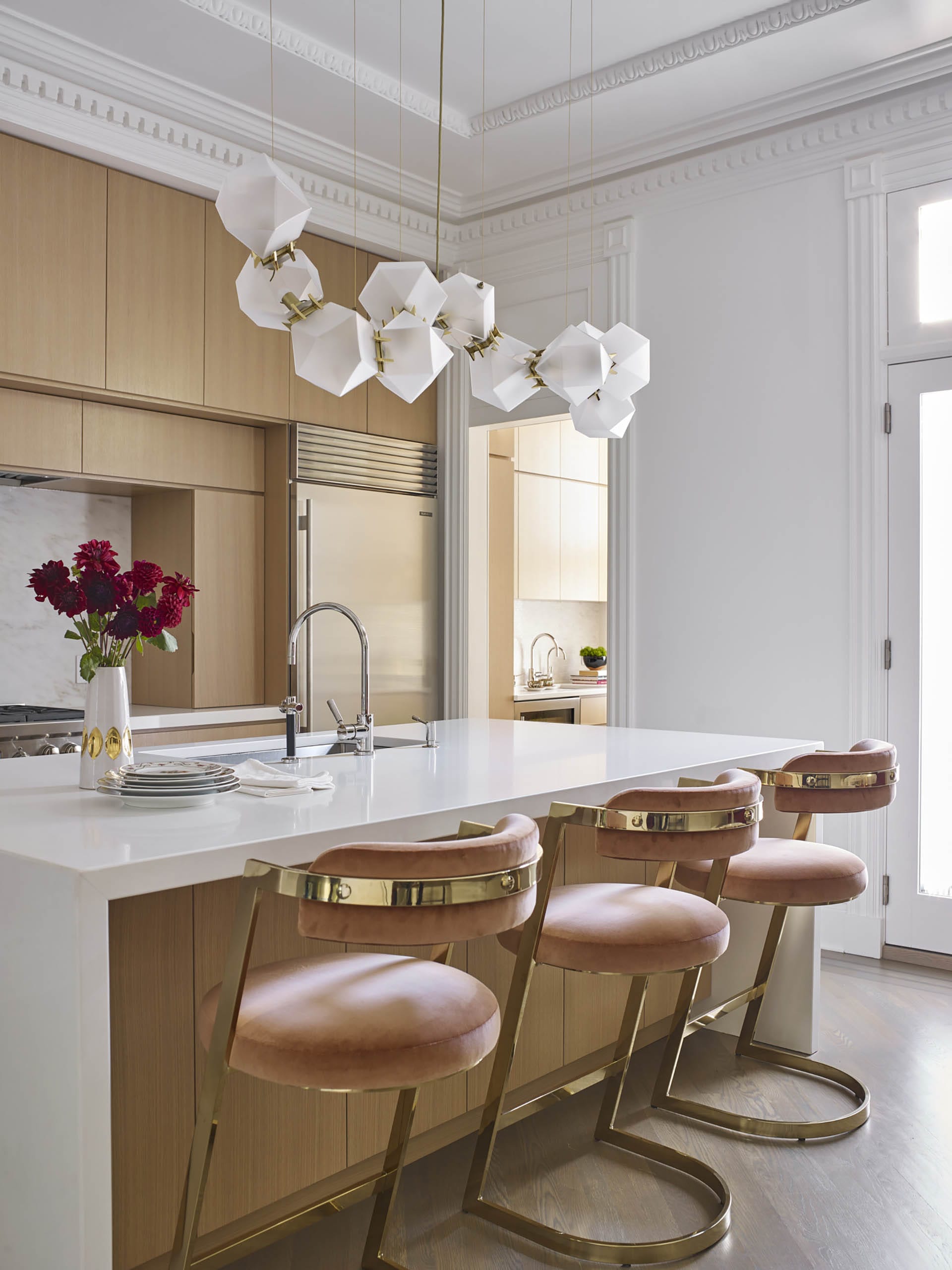 Kitchen with natural wood cabinetry, a white island and marble backsplash, restored historic crown molding, and three pink barstools at the island.