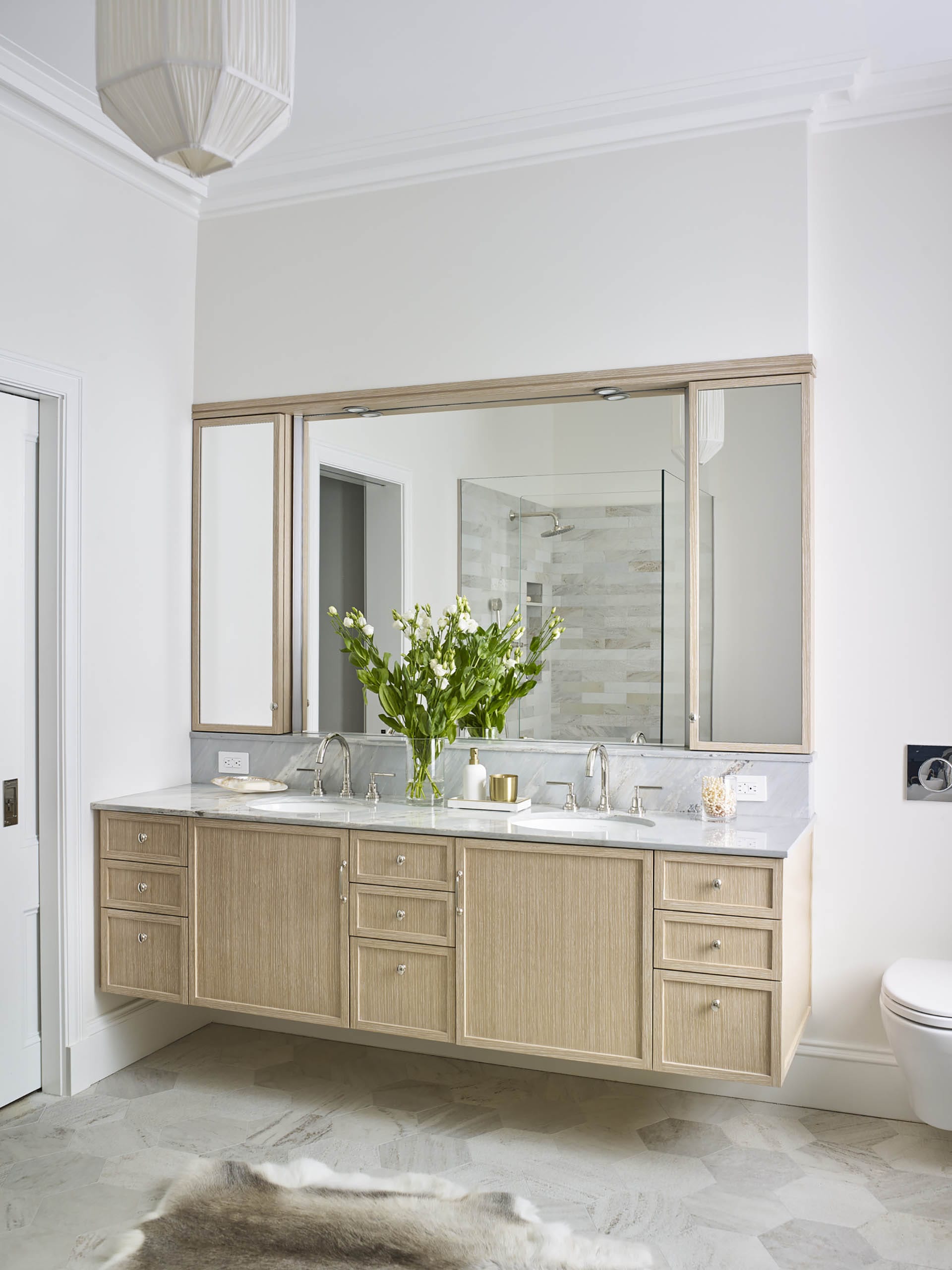 Natural wood vanity in a white primary bathroom with a vase of white flowers on the counter