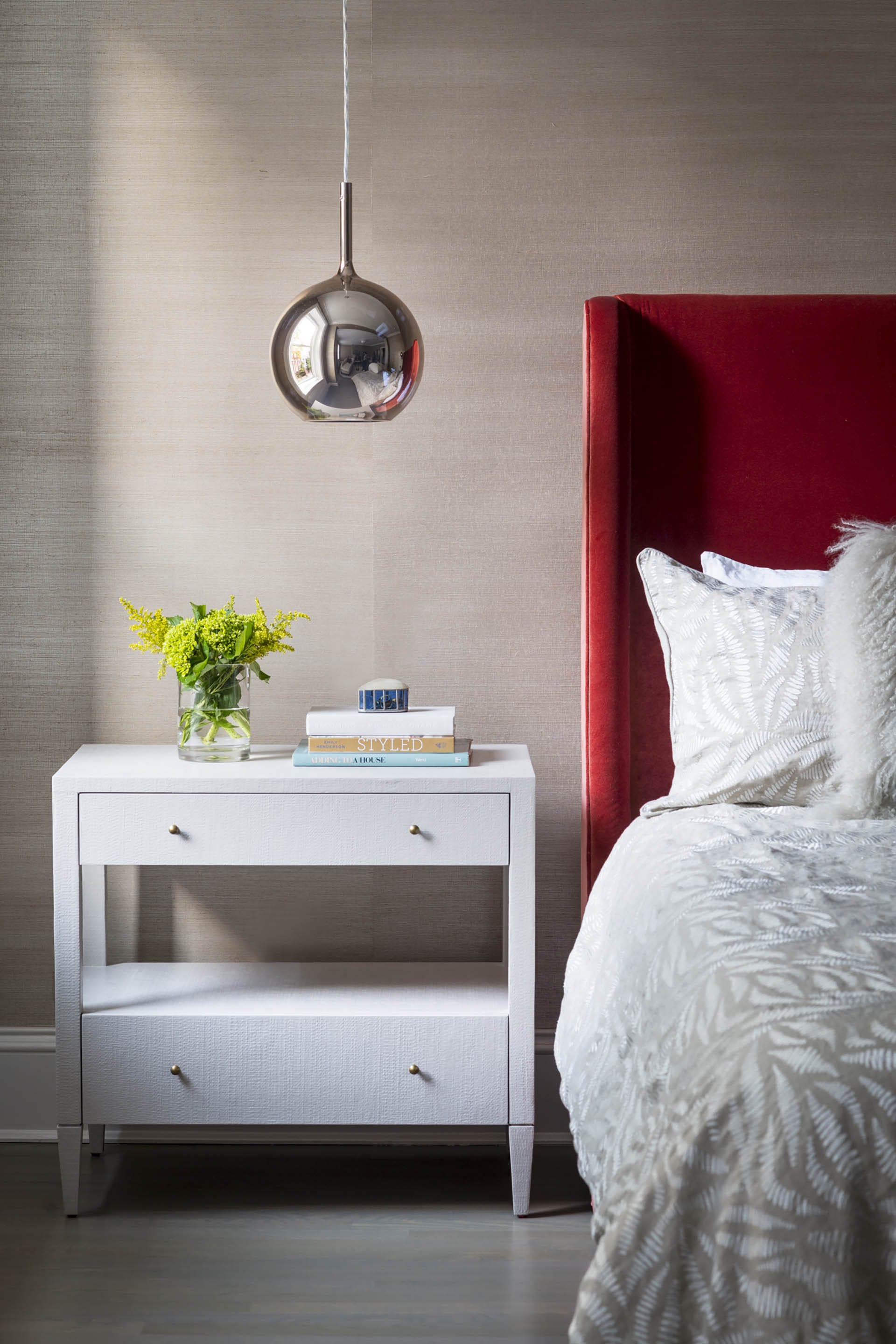 Primary bedroom with a rose-colored pendant light, grass cloth wallpaper, and red velvet upholstered headboard.