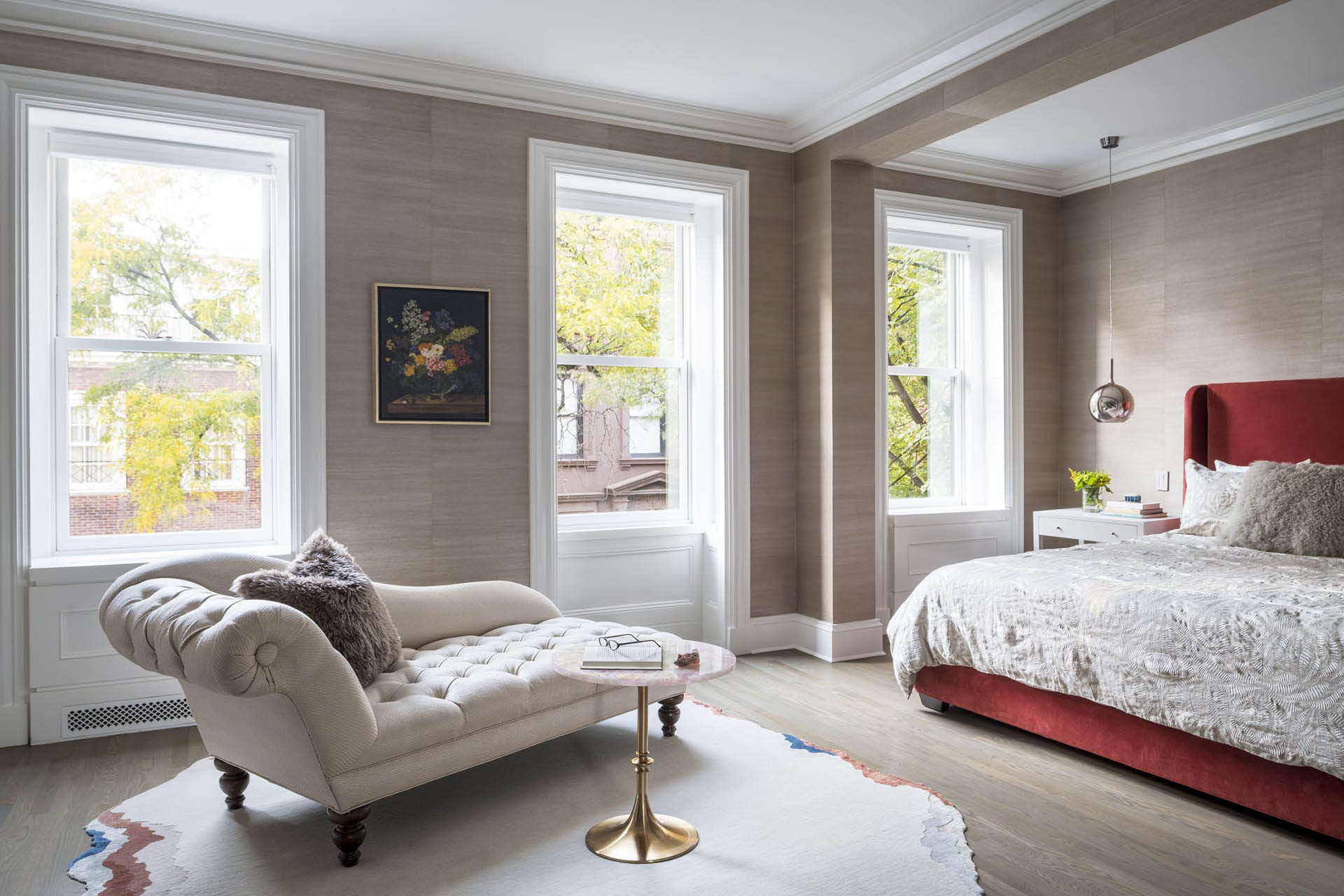 Primary bedroom with a tan settee, red velvet bed, and light-colored grass cloth wallpaper.