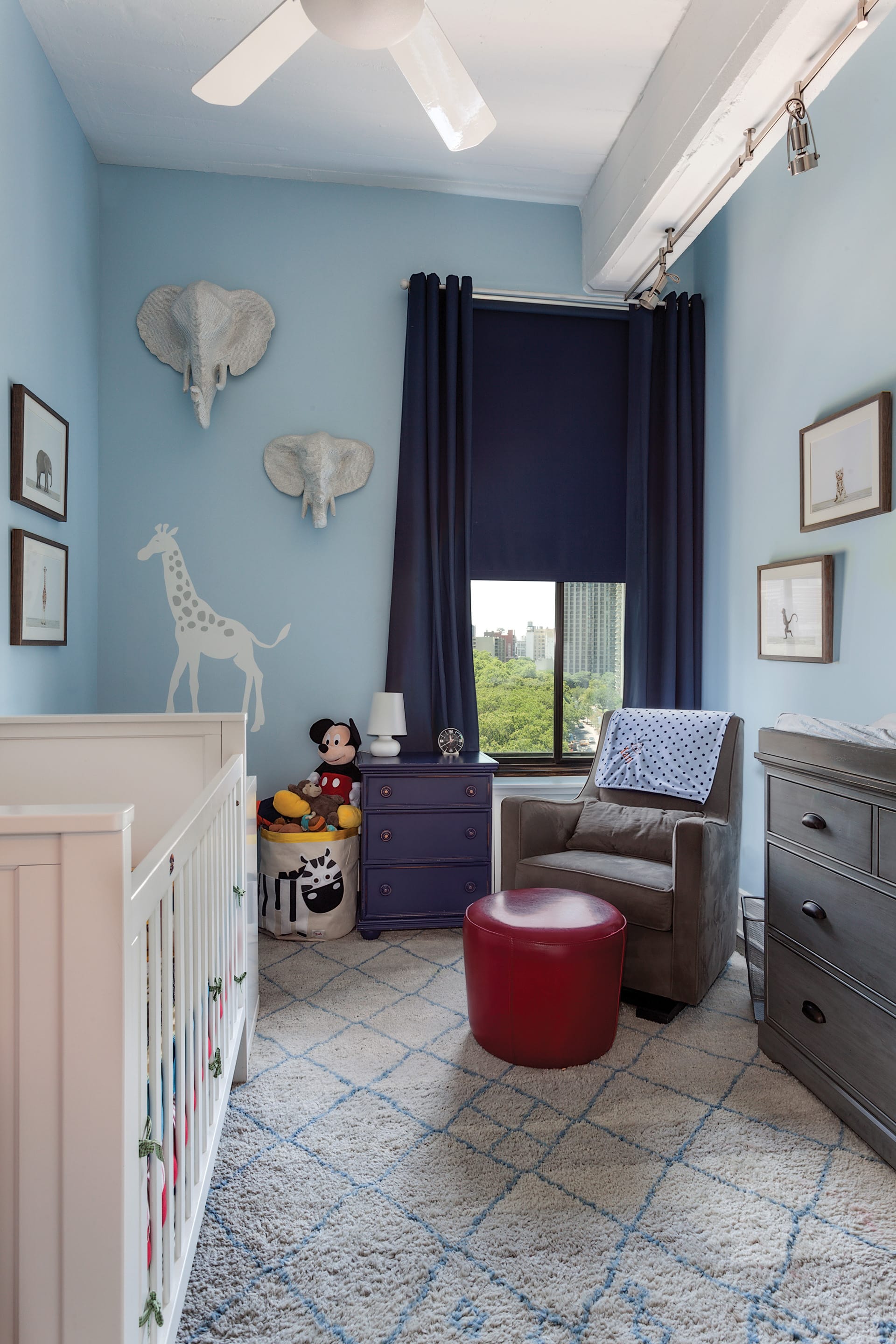 Nursery with a white crib, grey wood dresser, light blue walls, white animals on the wall, and navy blue curtains.