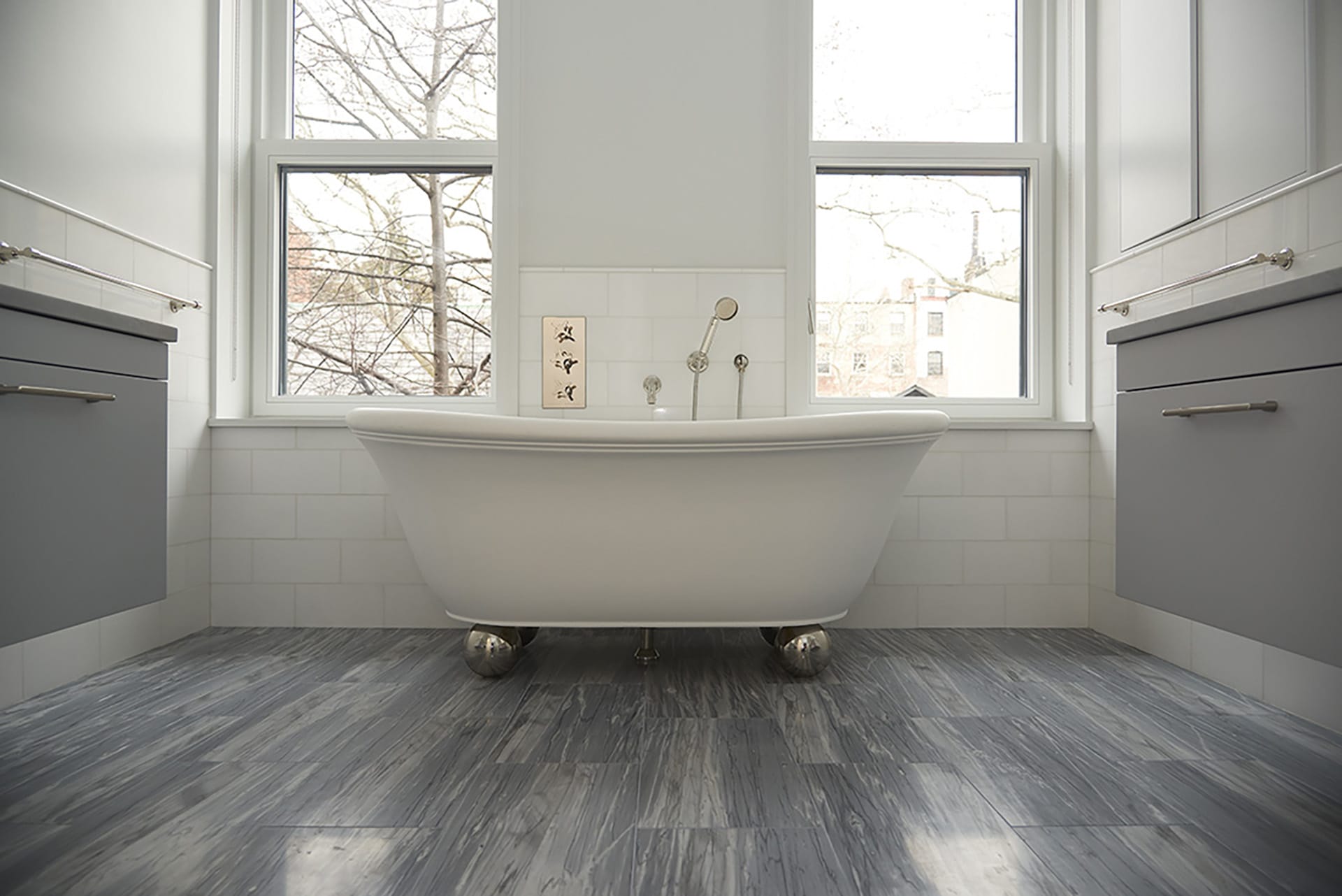 White freestanding tub next to two double hung windows in a primary bathroom suite with grey wood floors and cabinetry.