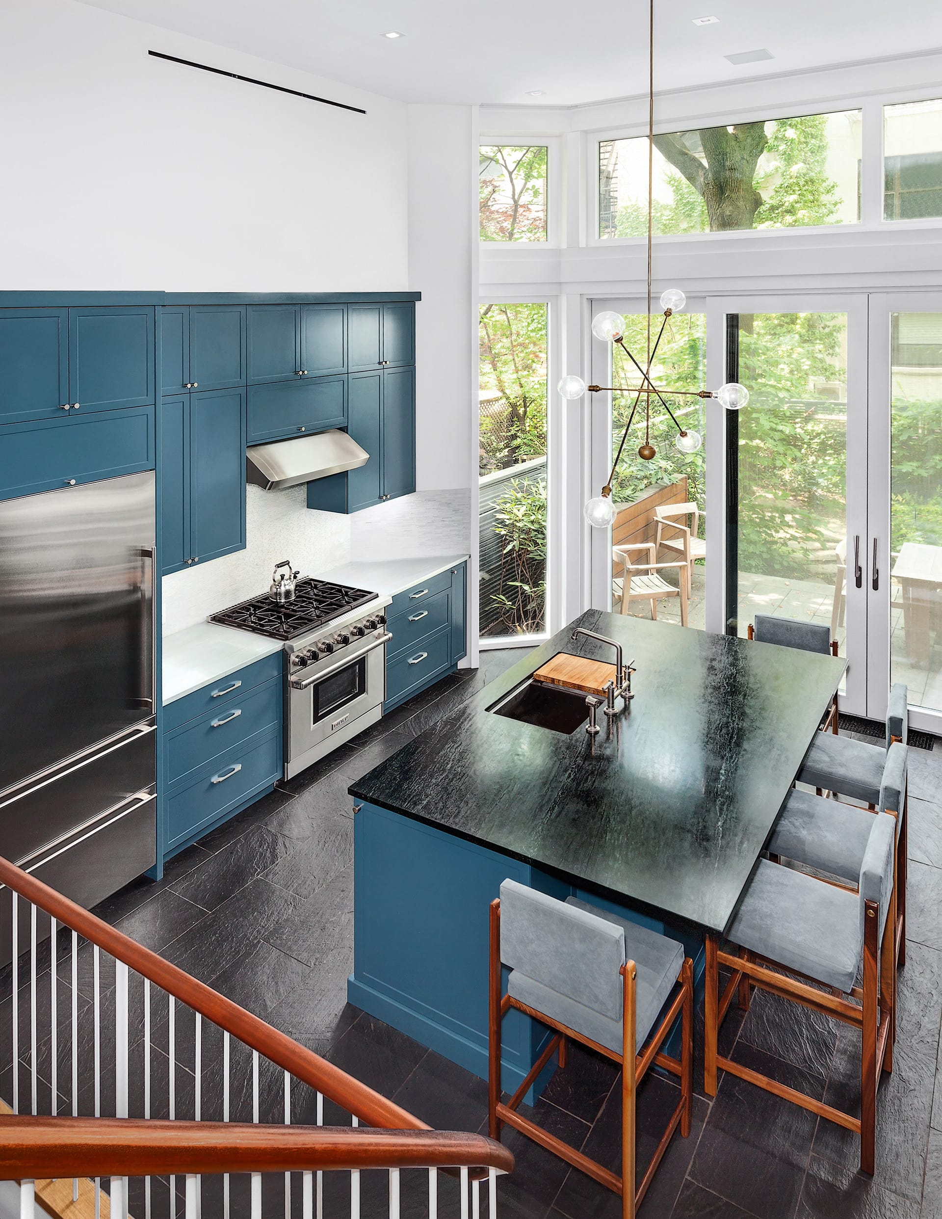 Double-height kitchen with teal cabinetry and island, wood and grey bar stools, and a large chandelier in front of large glass exterior doors