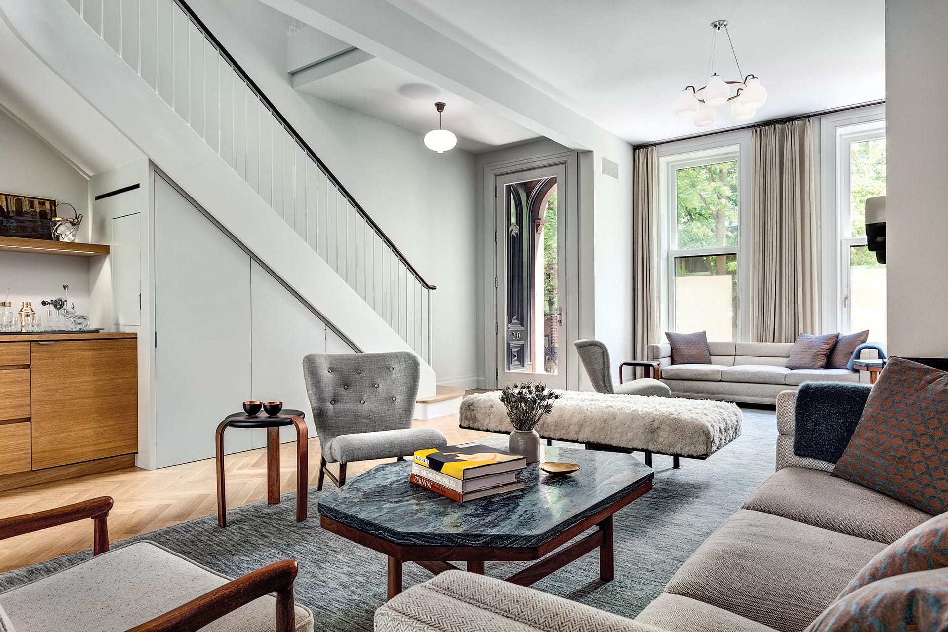 Living room and entry of a Brooklyn Heights Passive House. A dry bar sits underneath the staircase in front of the front door. Chairs, a coffee table, and couches are atop a grey carpet.