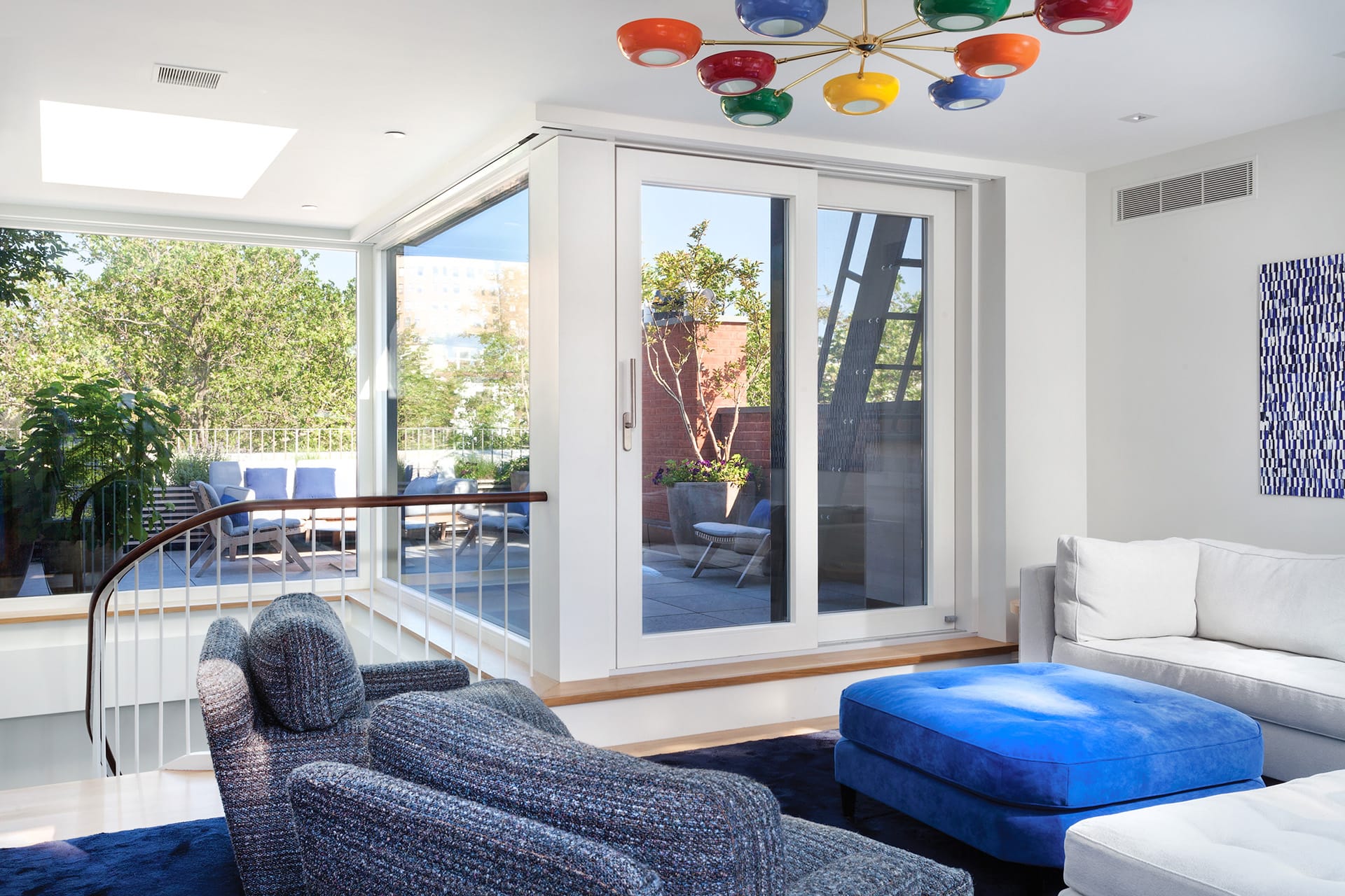 Penthouse in a Brooklyn Heights townhouse with blue carpet, painting, ottoman, and armchairs. A colorful light fixture hangs off the ceiling, and the rooftop deck can be seen through the large windows and glass door.