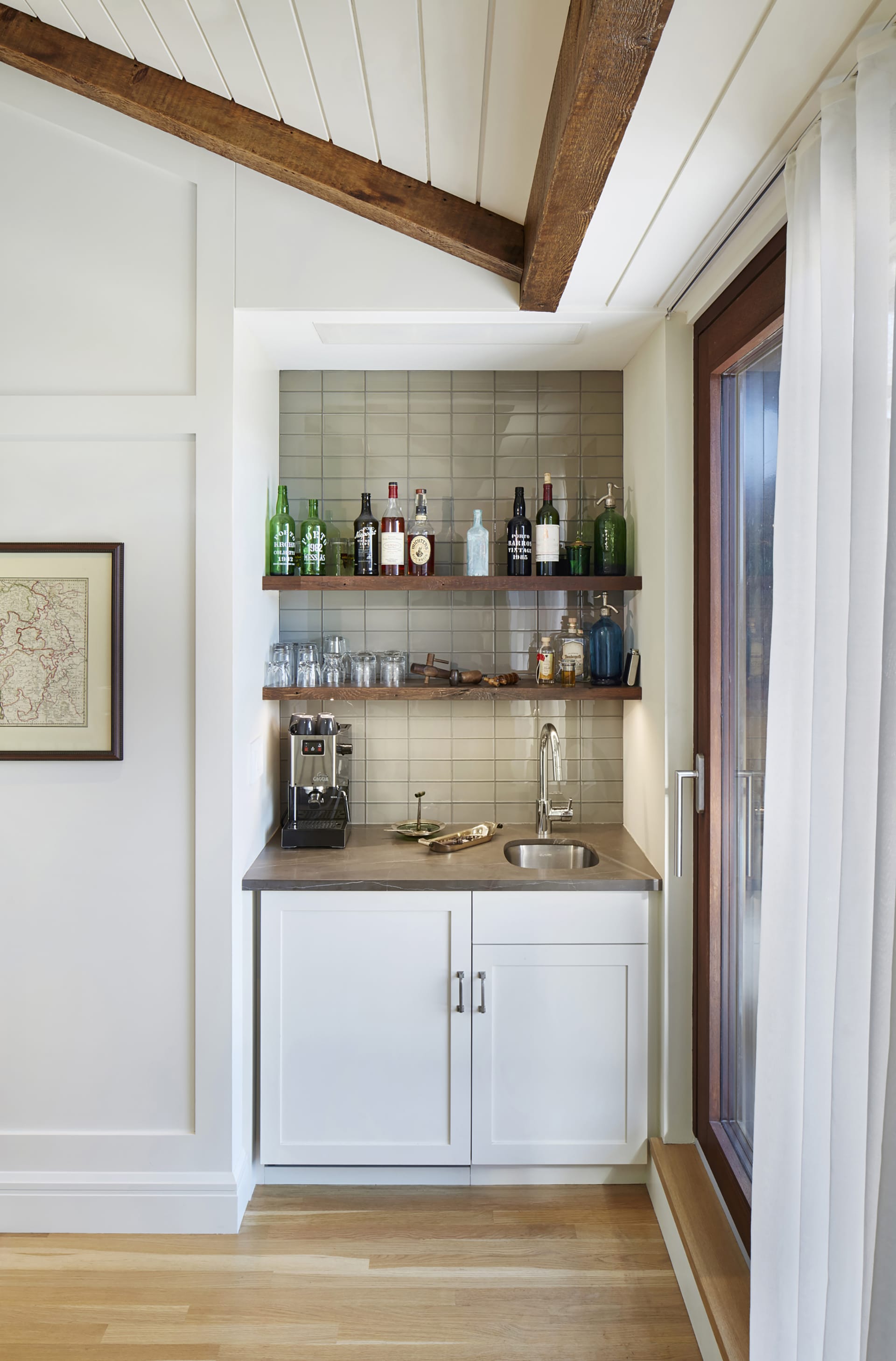 Wet bar with tan backsplash, wood shelves, and a grey countertop. The ceiling of the room is angular and accented by exposed wood beams.