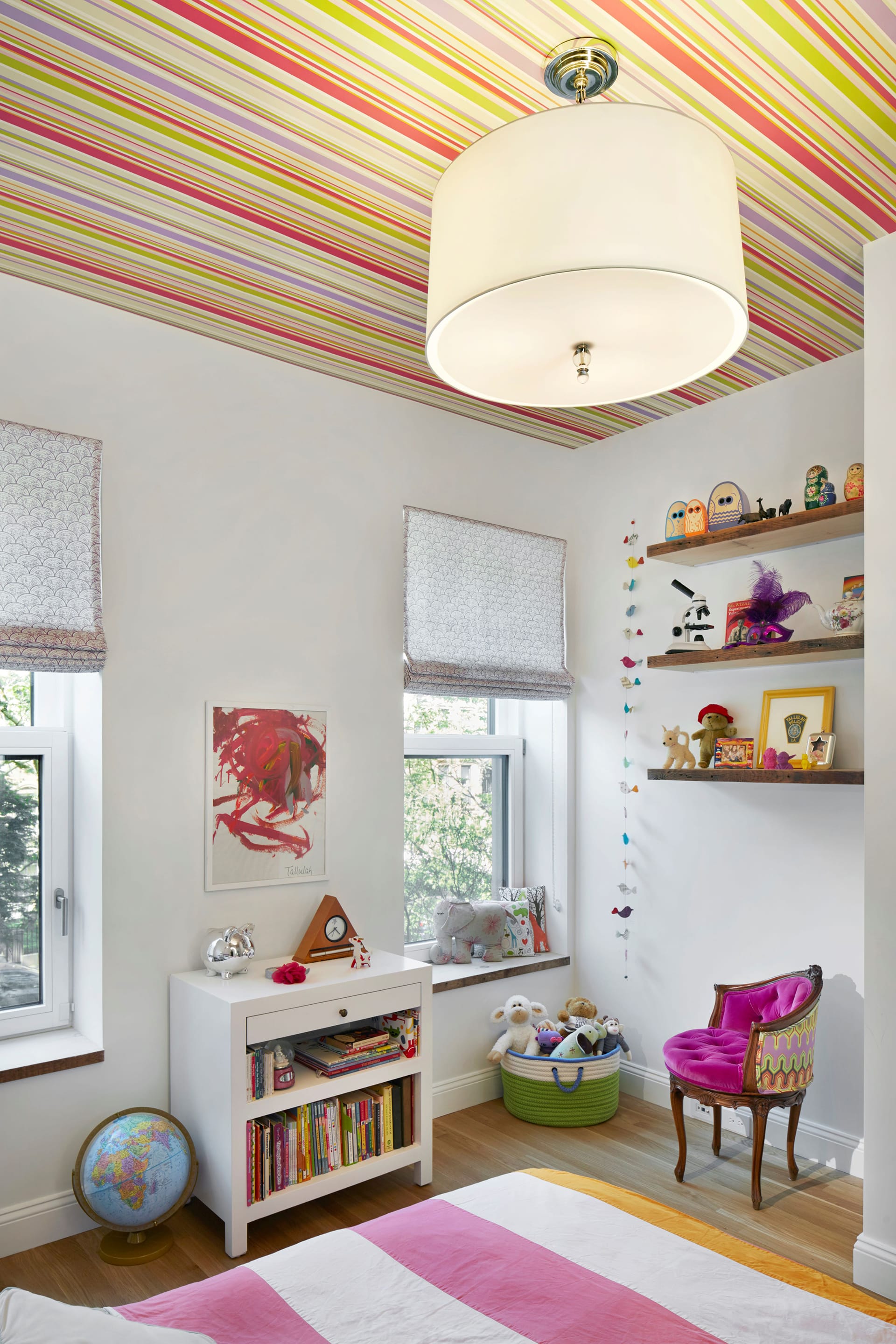 Corner of a child's bedroom with striped wallpaper on the ceiling, suspended bookshelves, Roman shades, and a white and pink striped comforter at the bottom of the frame.