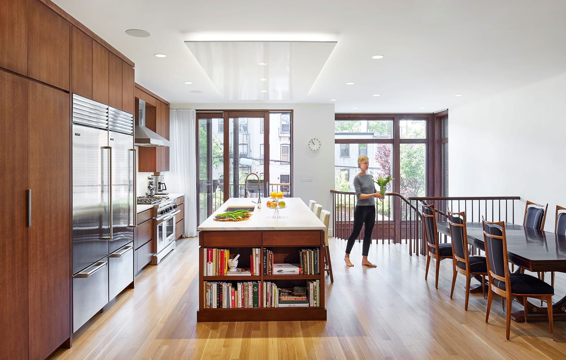 Eat-in kitchen with dark wood cabinetry and accents, a marble-topped island, and a sliding glass door leading to a juliet balcony. To the right of the image, a dark wood dining table is in front of a staircase leading down to the next level of the house. A blonde woman holding a vase of flowers walks from the left to the right of the frame.