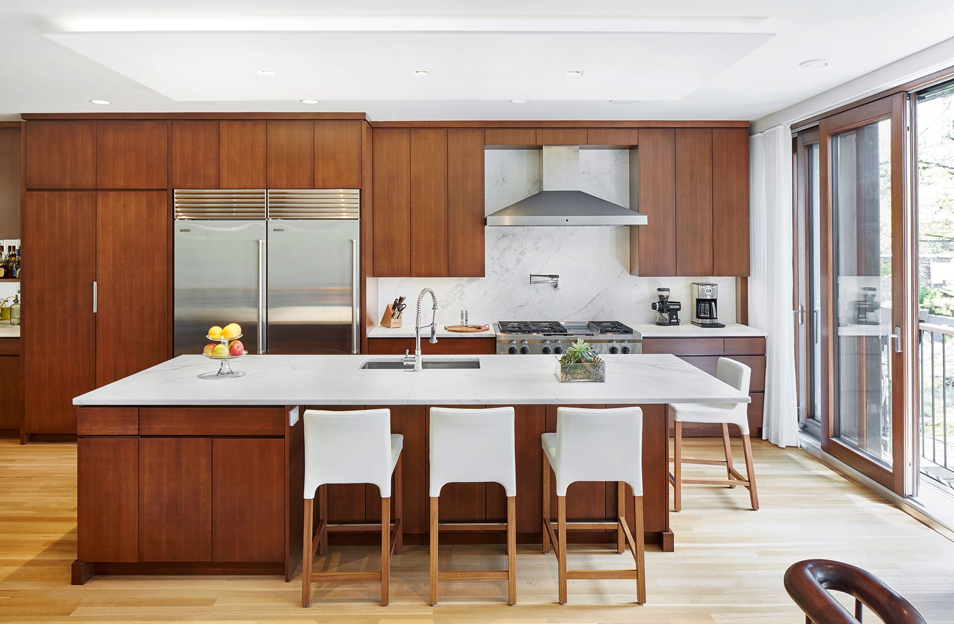 A kitchen with dark wood cabinetry, a metal range hood and refrigerator, marble-topped cherry island and marble backsplash.