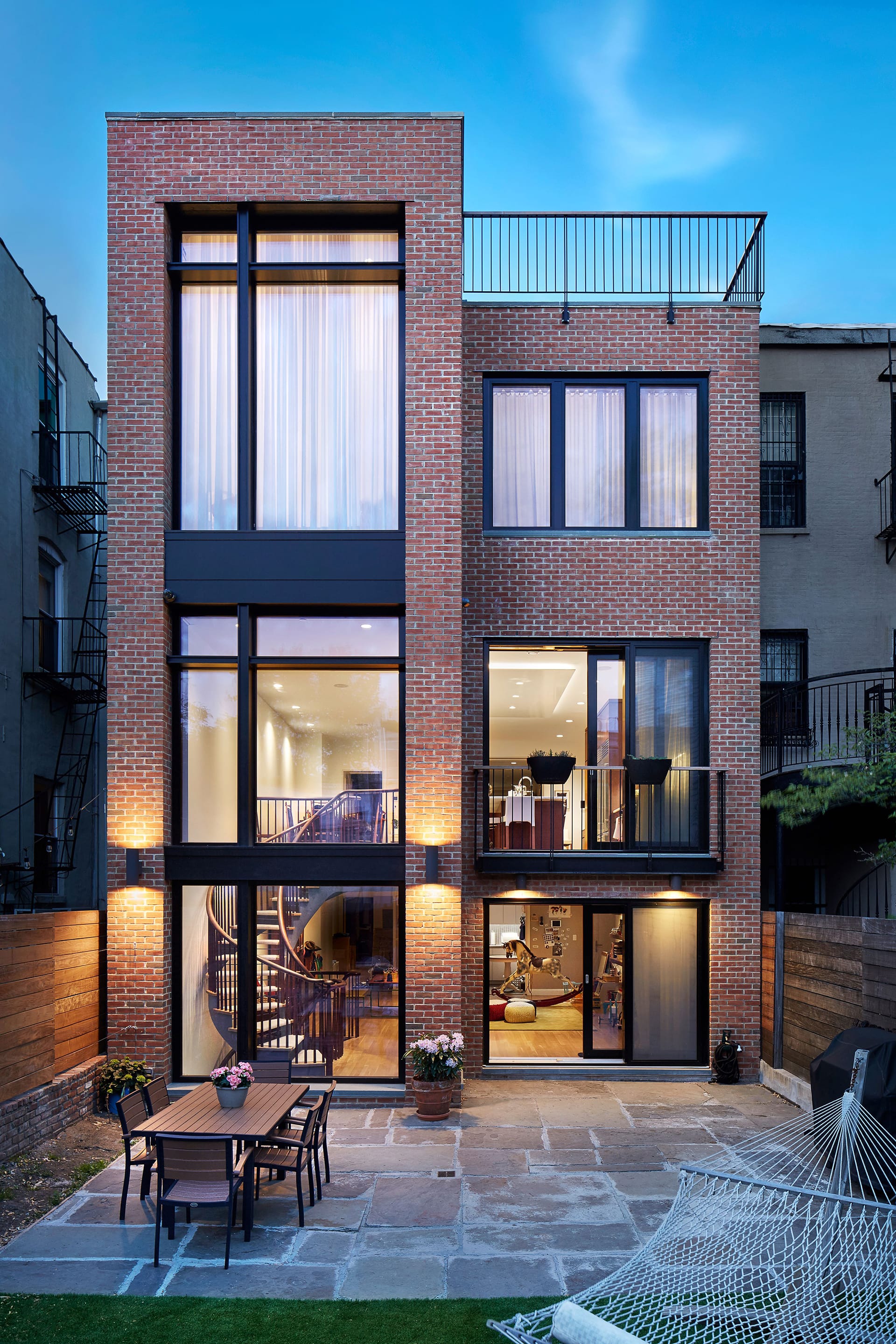 Rear facade of a Carroll Gardens Passive House with three stories of glass on the left side, and large expanses of glass at the right. Atop the right side of the building, railing creates a barrier for the roofdeck. Stone pavers separate the grass from a dining area with a hammock in the back yard.