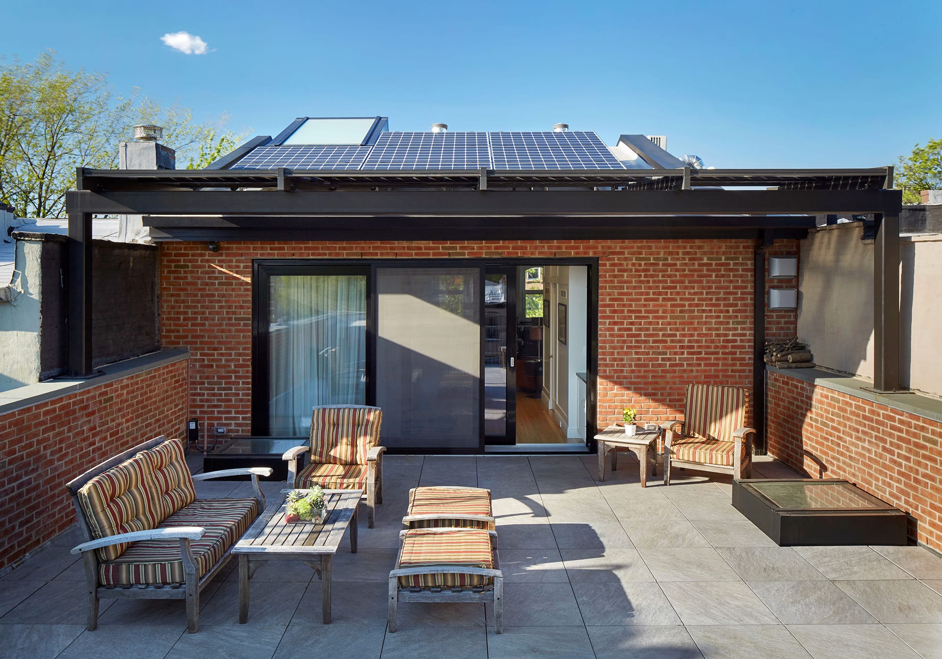 Penthouse of a Carroll Gardens townhouse with bluestone pavers, striped outdoor furniture, sliding glass doors, and a rooftop solar array.