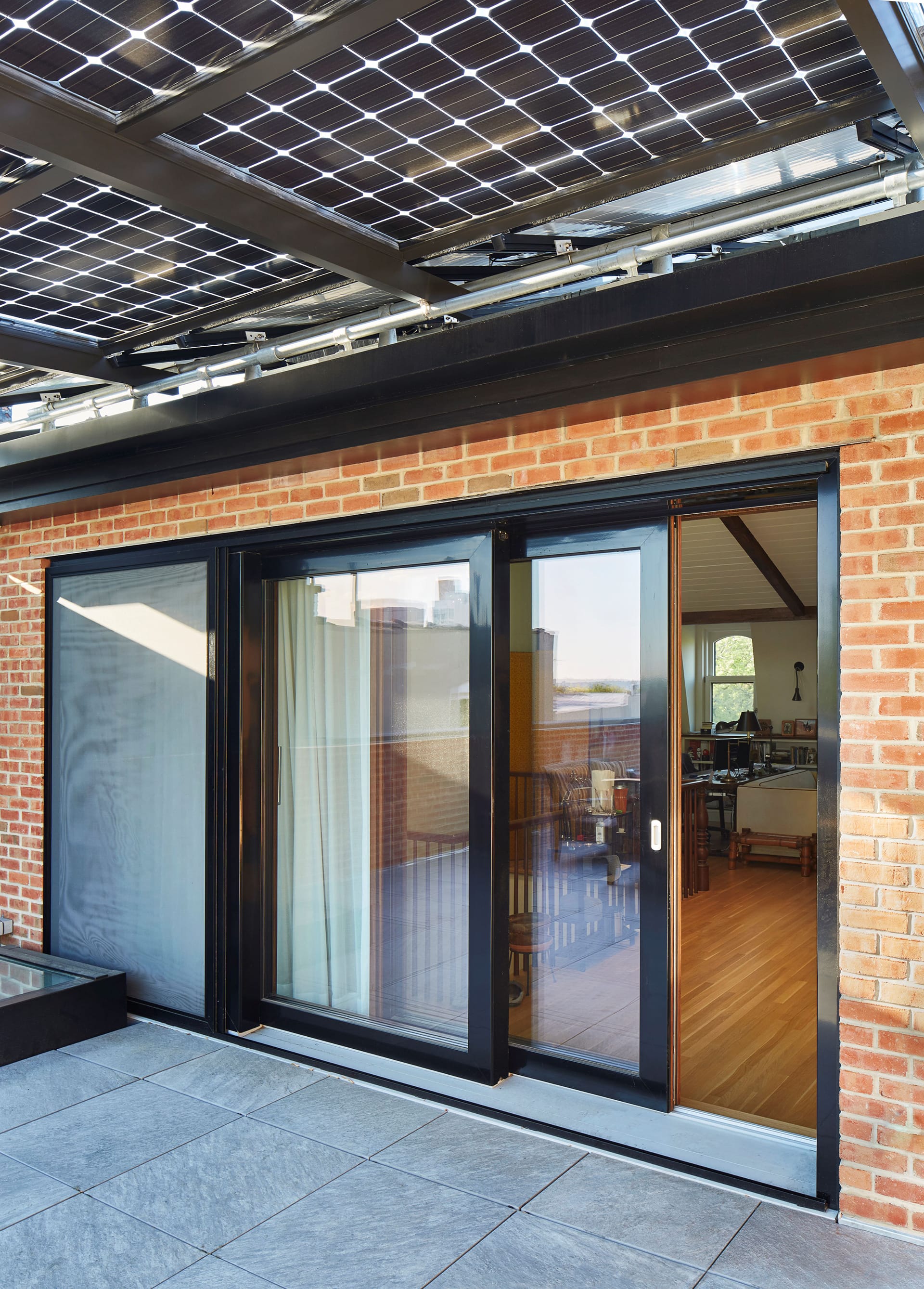 Large sliding glass doors separating an attic space from the roof deck. The roof deck has bluestone pavers and is covered by a solar canopy.