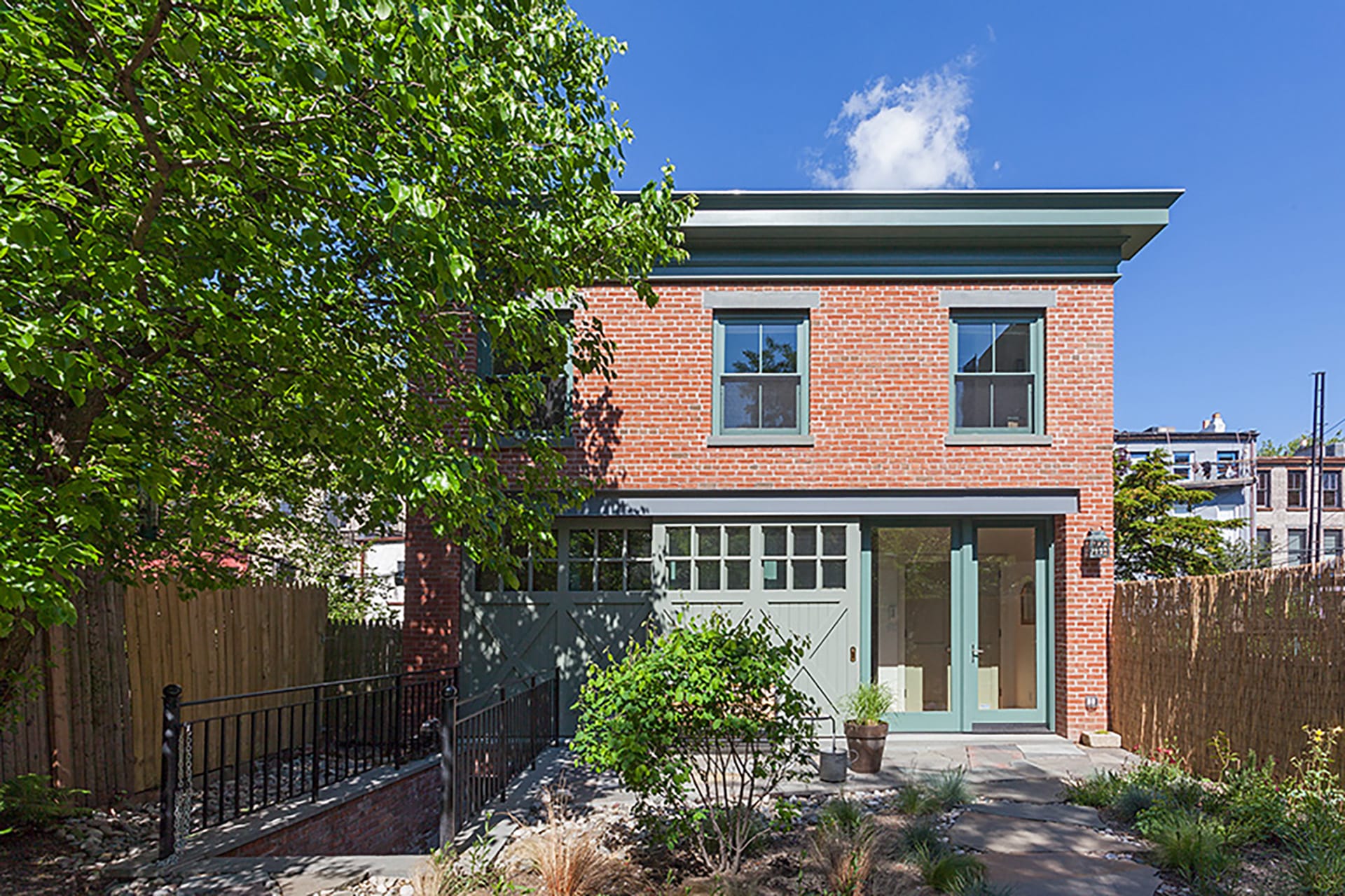 Front facade of a freestanding brick carriage house with large barn doors, green trim, a staircase leading to the basement entrance, and a front garden