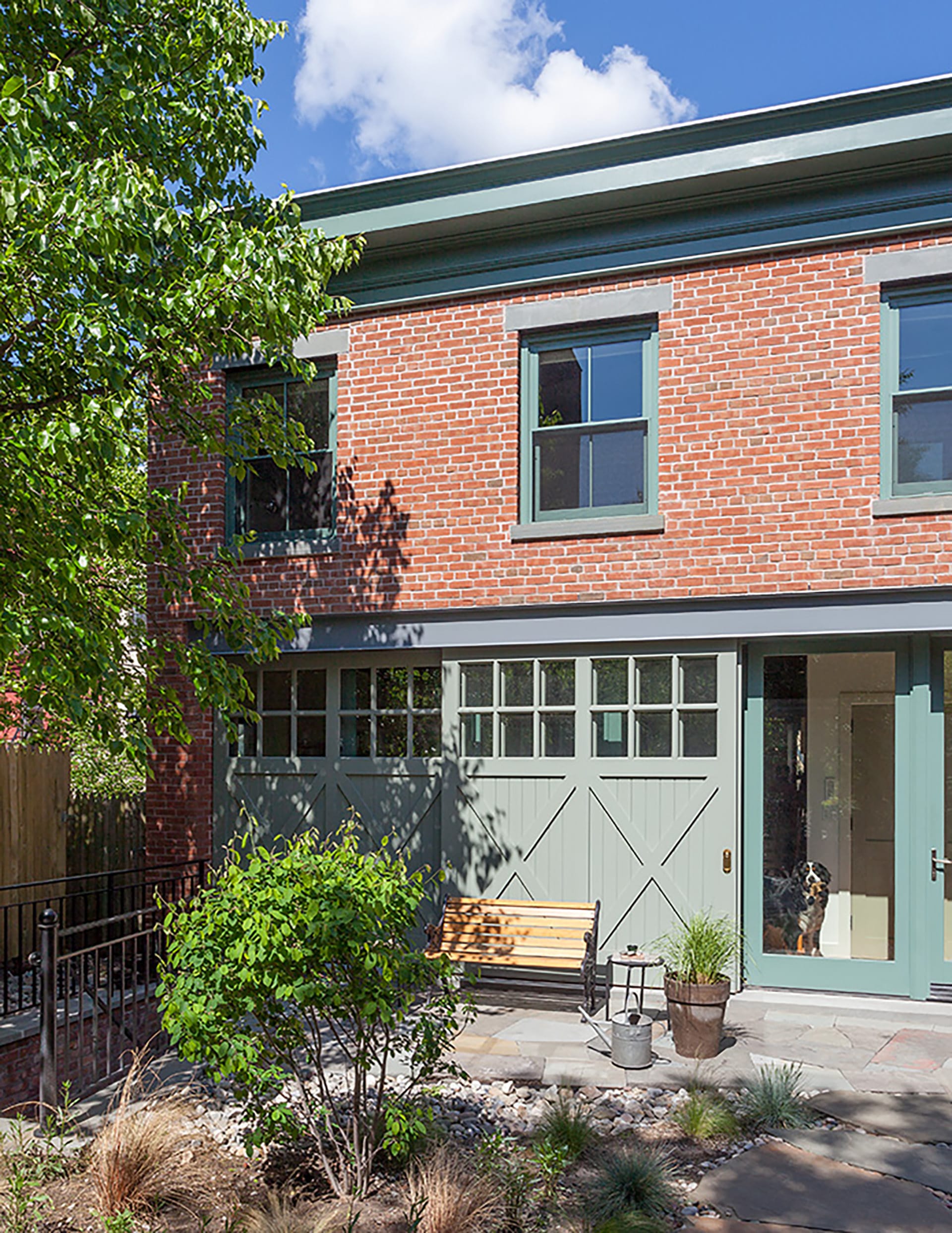 Front facade of a freestanding brick carriage house with large barn doors, green trim, a staircase leading to the basement entrance, and a front garden