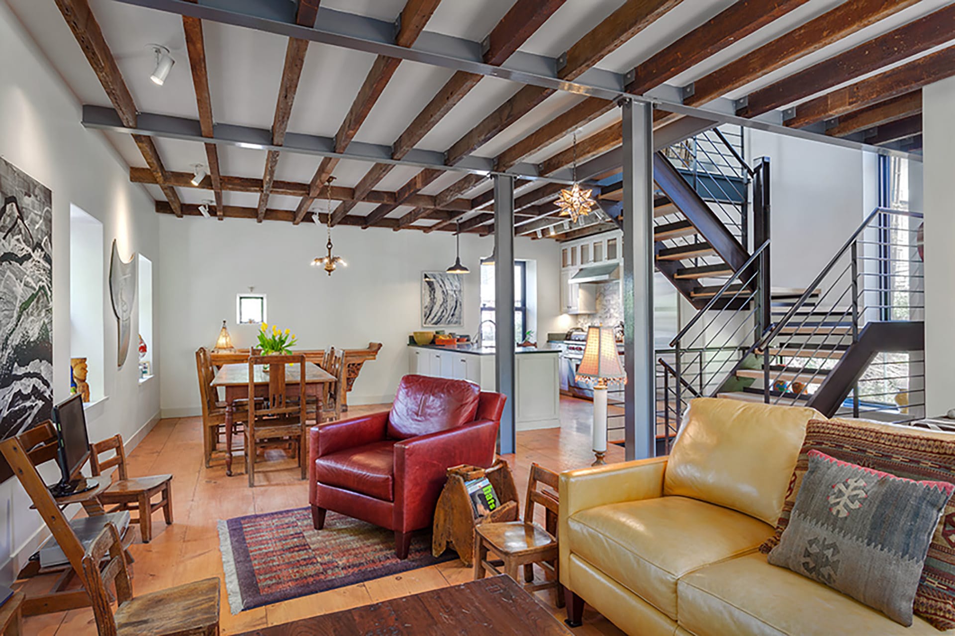 Living space with exposed ceiling beams, a yellow leather couch and red leather armchair, dining area, and modern metal staircase in a Cobble Hill carriage house