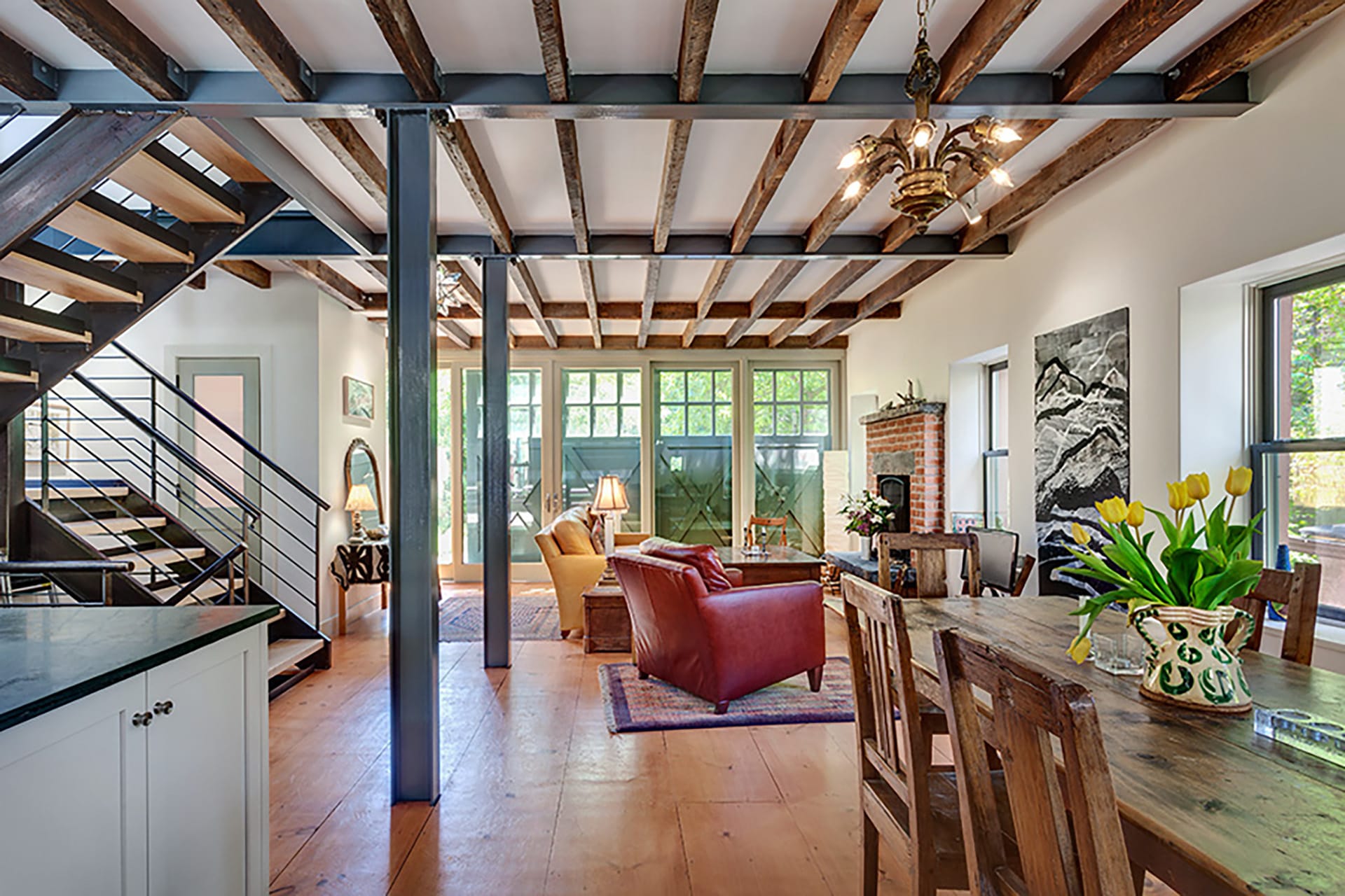 Living space with exposed ceiling beams, a yellow leather couch and red leather armchair, dining area, and modern metal staircase in a Cobble Hill carriage house