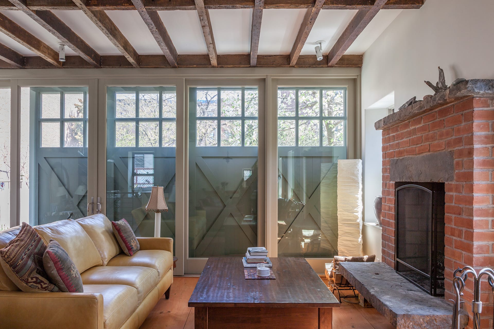 Living space in a carriage house with a wall of glass doors covered by sliding barn doors, exposed beams, original fireplace, and yellow leather couch.
