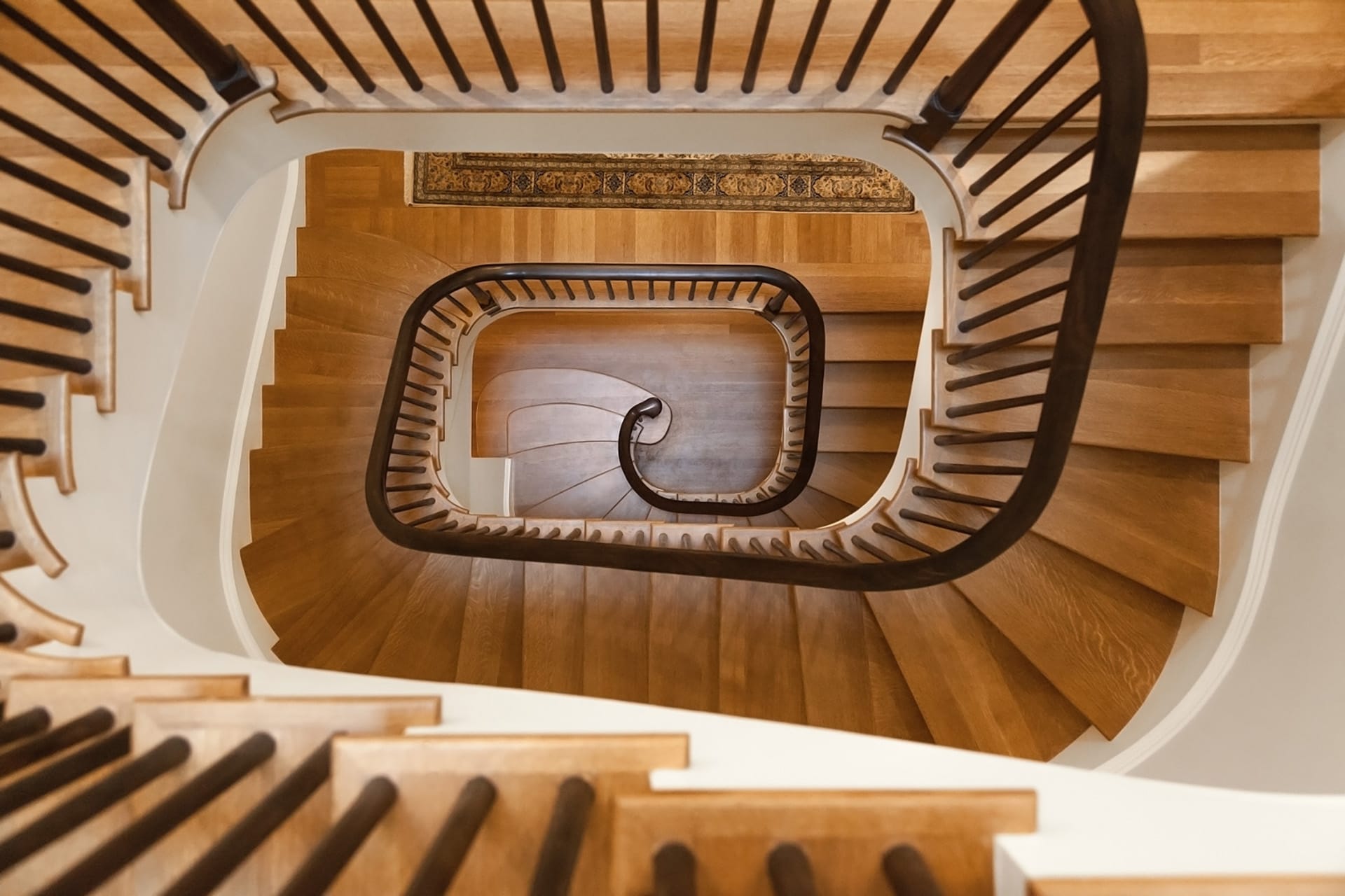 Looking down from the third story of a sculptural staircase with dark wood railings and lighter wood treads.