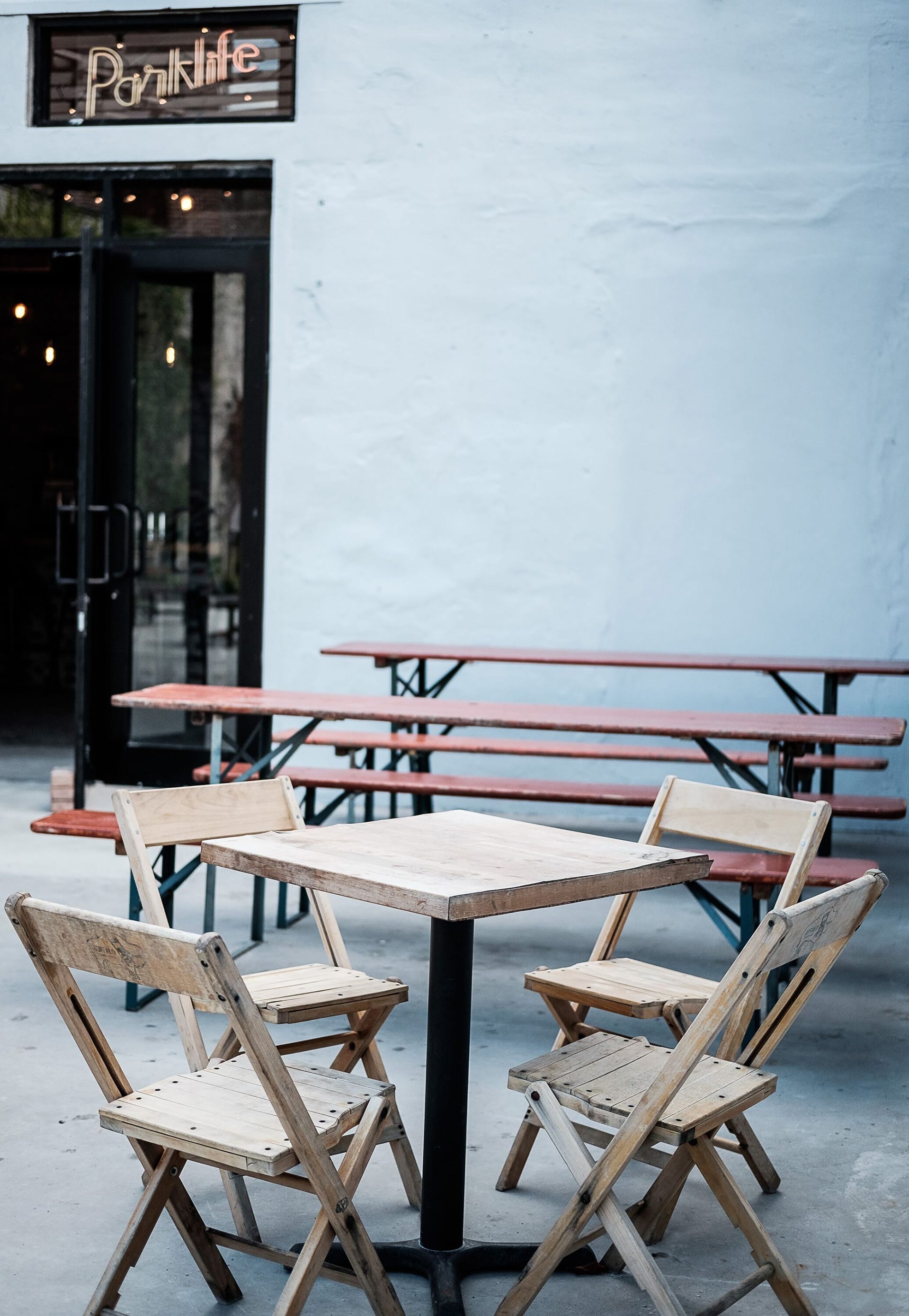 Parklife outdoor seating areas with a table in the foreground and two picnic tables in the background. The exterior wall is painted light grey and a neon Parklife sign above the door.