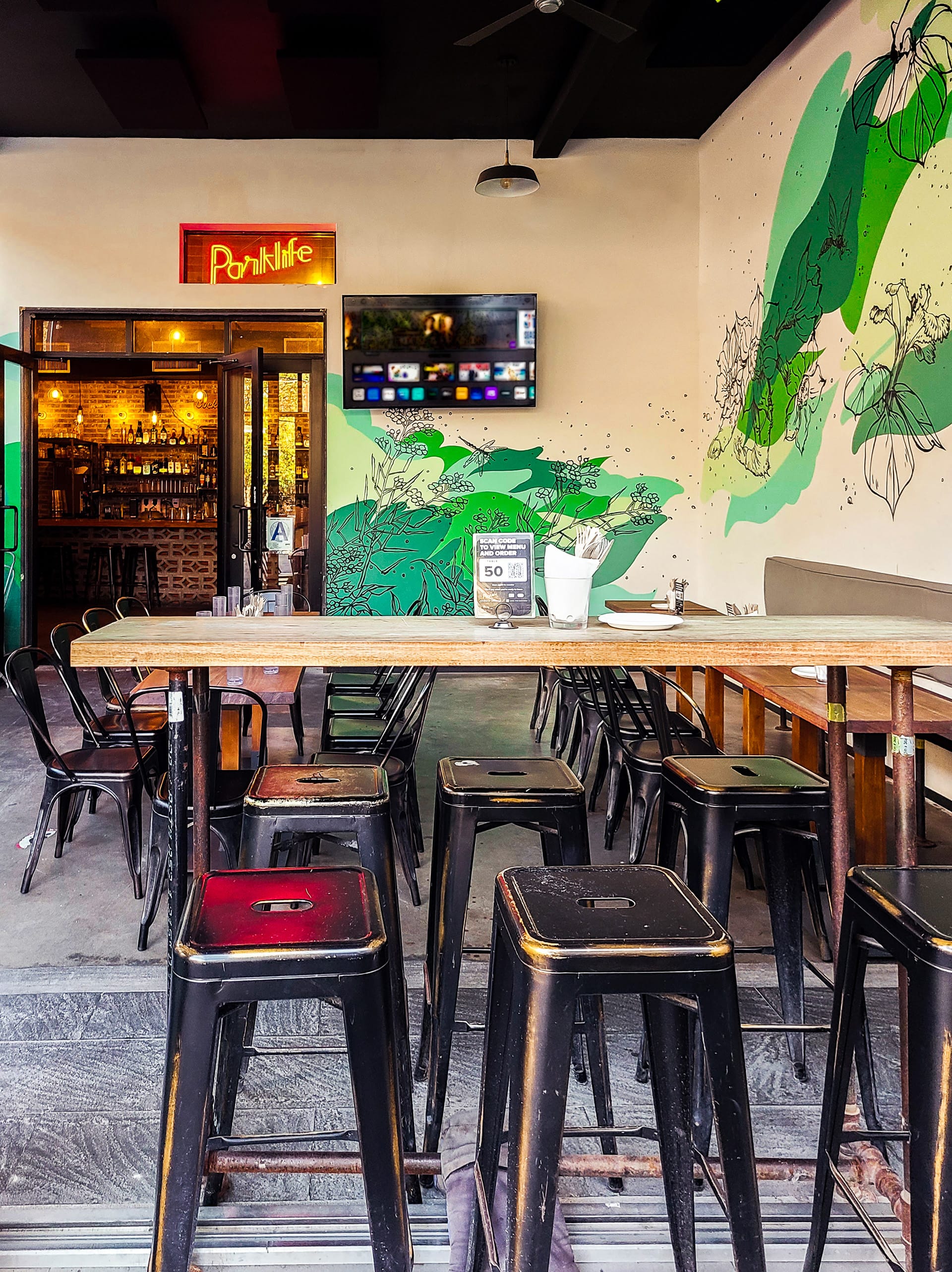 Entrance to Parklife with a green mural wrapping around the walls, a neon Parklife sign, wood bar, black barstools, and black chairs.