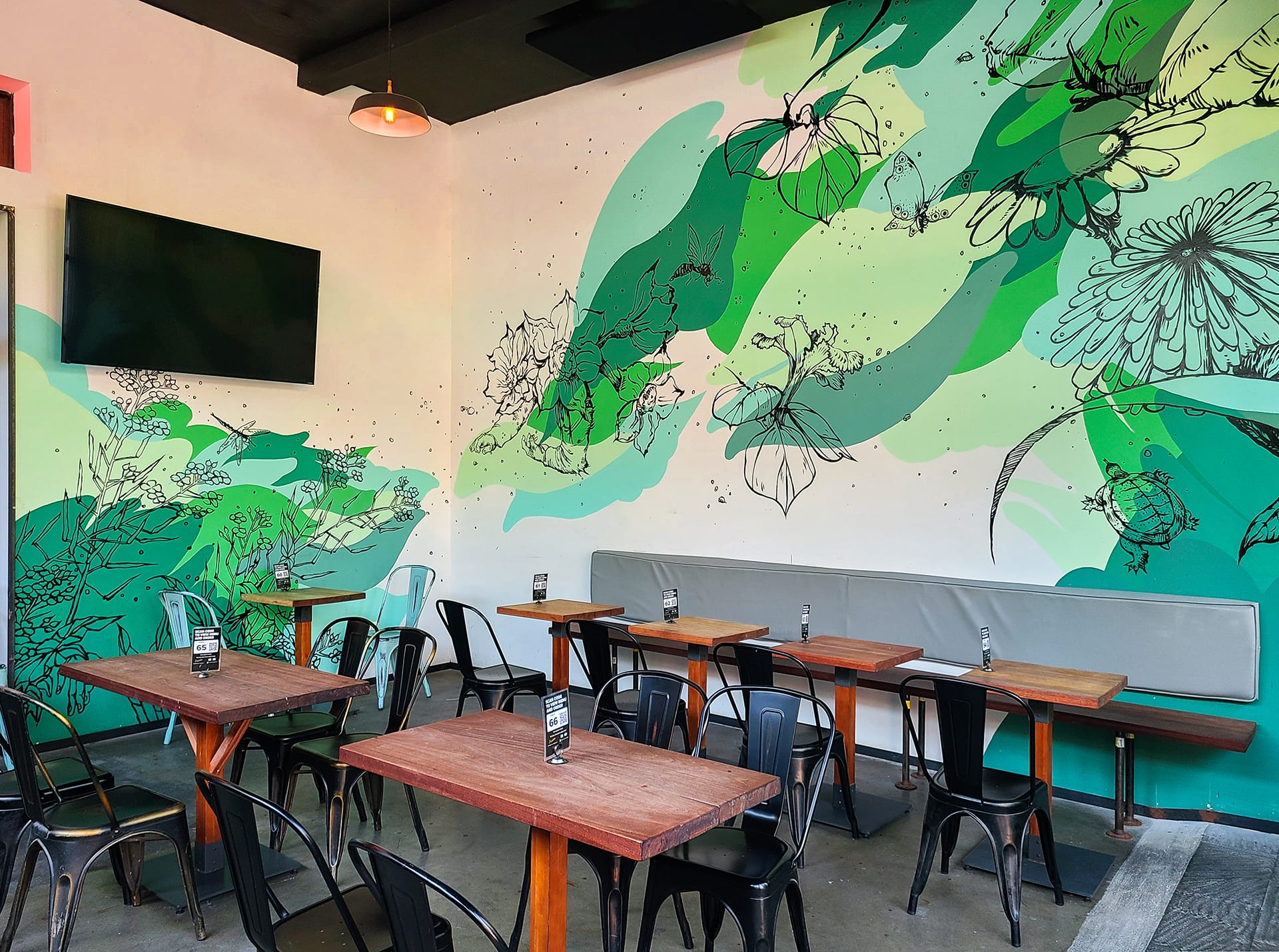 Outdoor seating area with a green floral mural, wood tables, and black chairs.