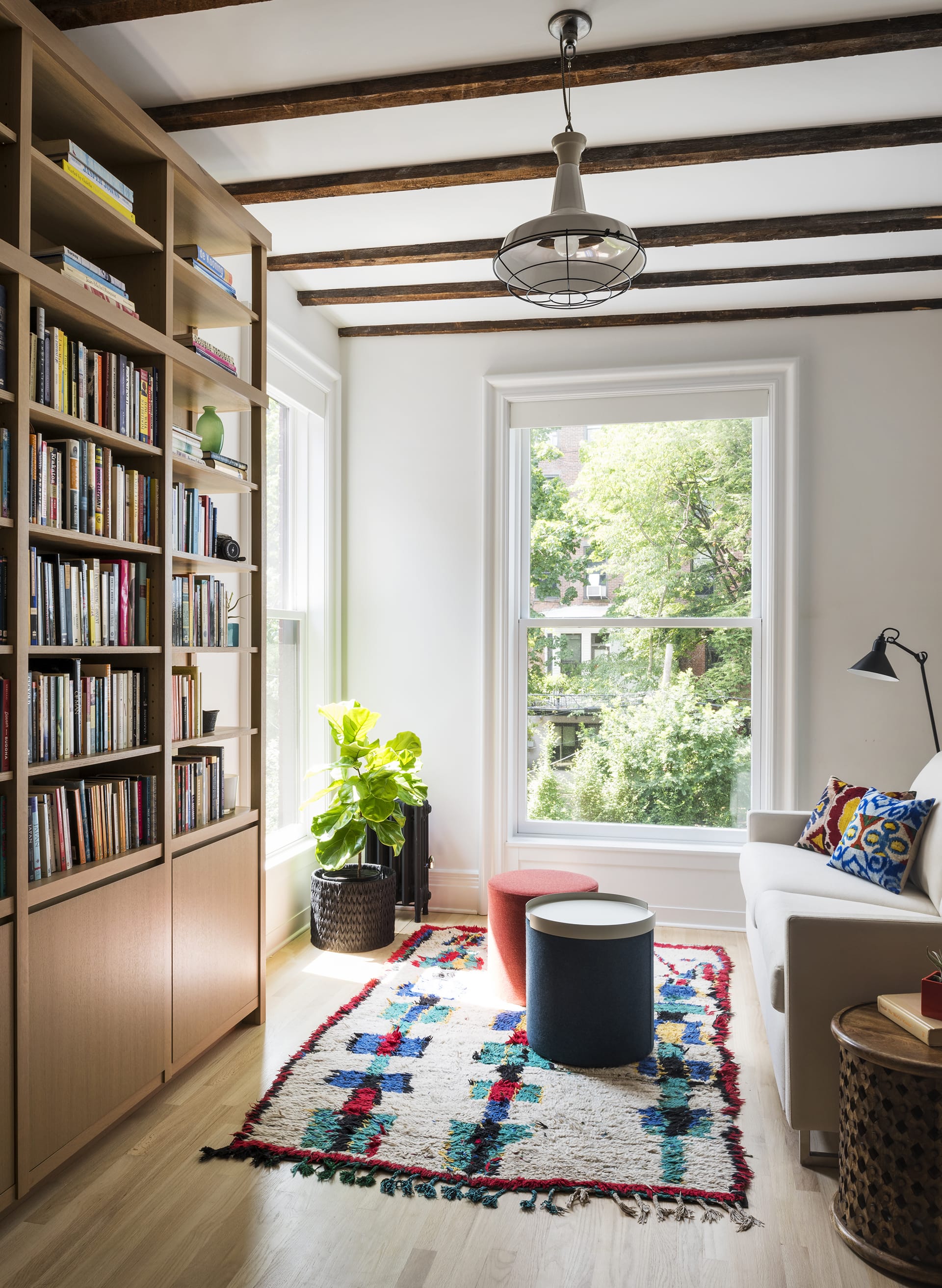 Reading room with a tan couch across from a large wood bookshelf in a small room with exposed ceiling beams and bright carpet and pillows.