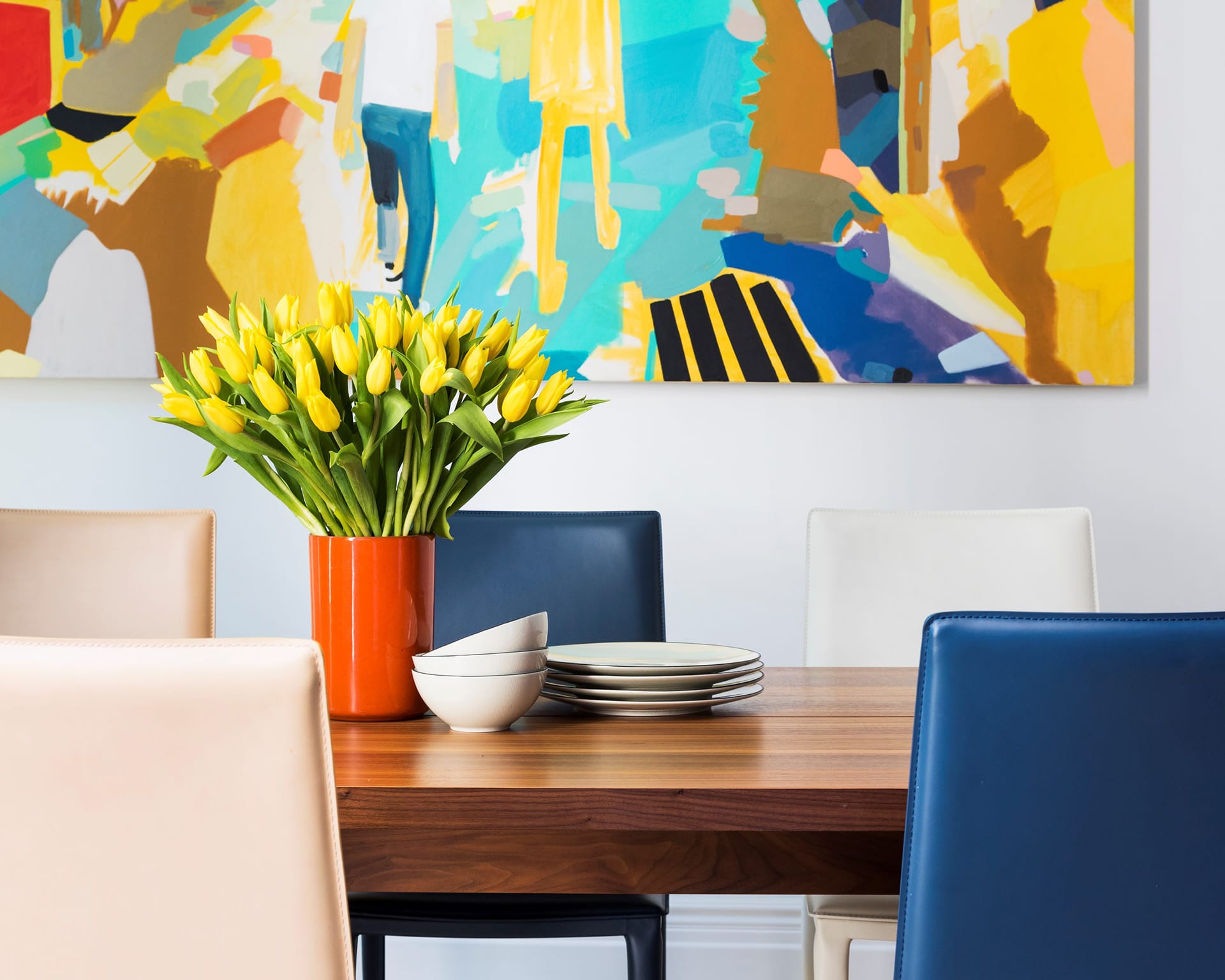 Dining room with mixed colored leather chairs, a wood table, and a large colorful painting on the wall.