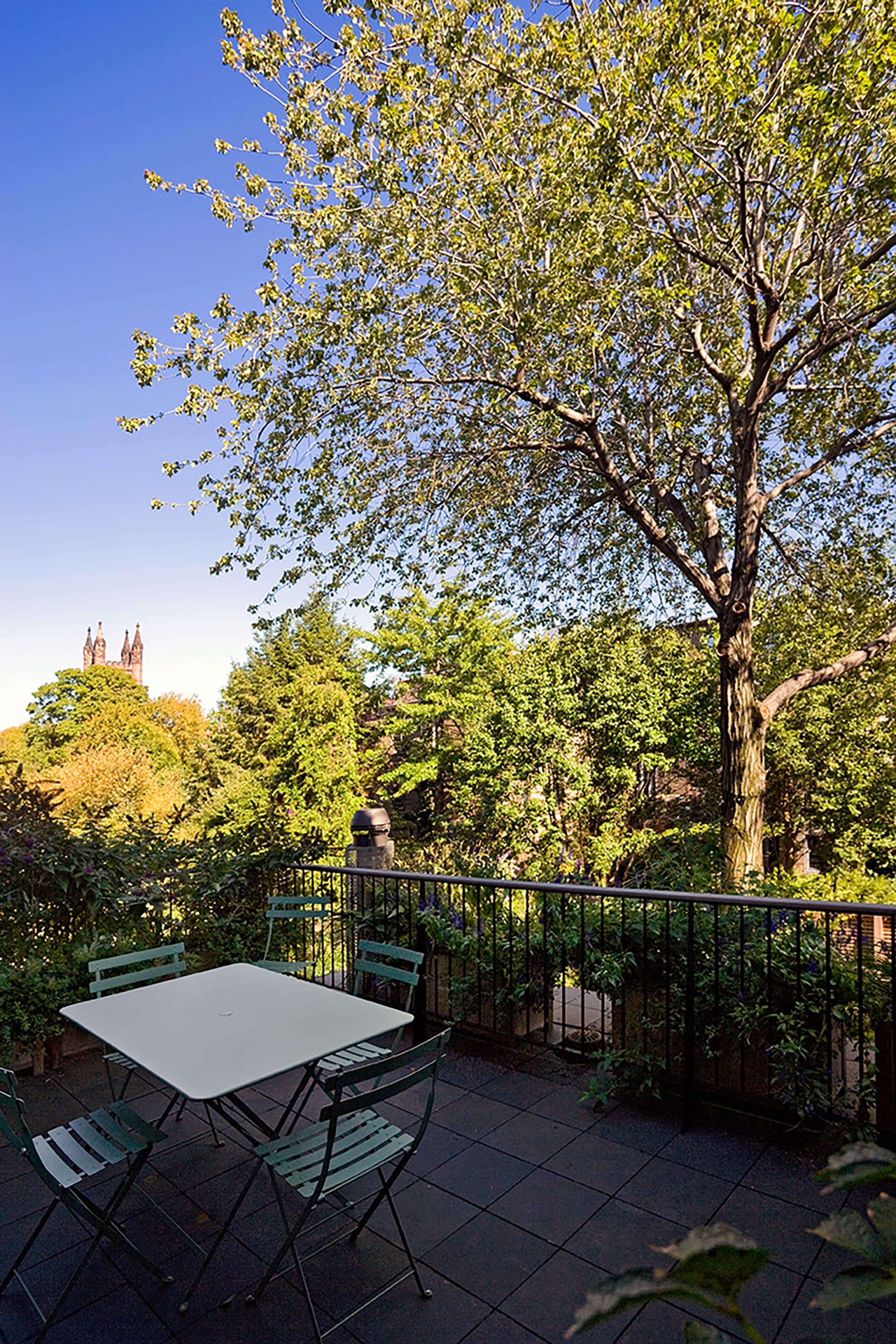 Terrace with stone pavers, a white table, and four green outdoor chairs looking at treetops.