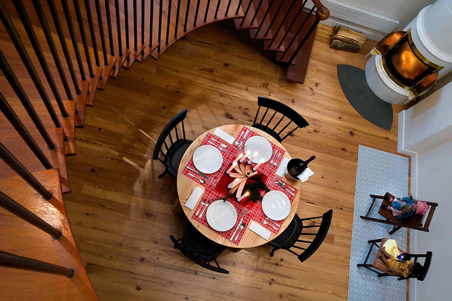 Circular kitchen table with black chairs, next to a white Scandinavian fireplace, in a double height space underneath a spiraling staircase.