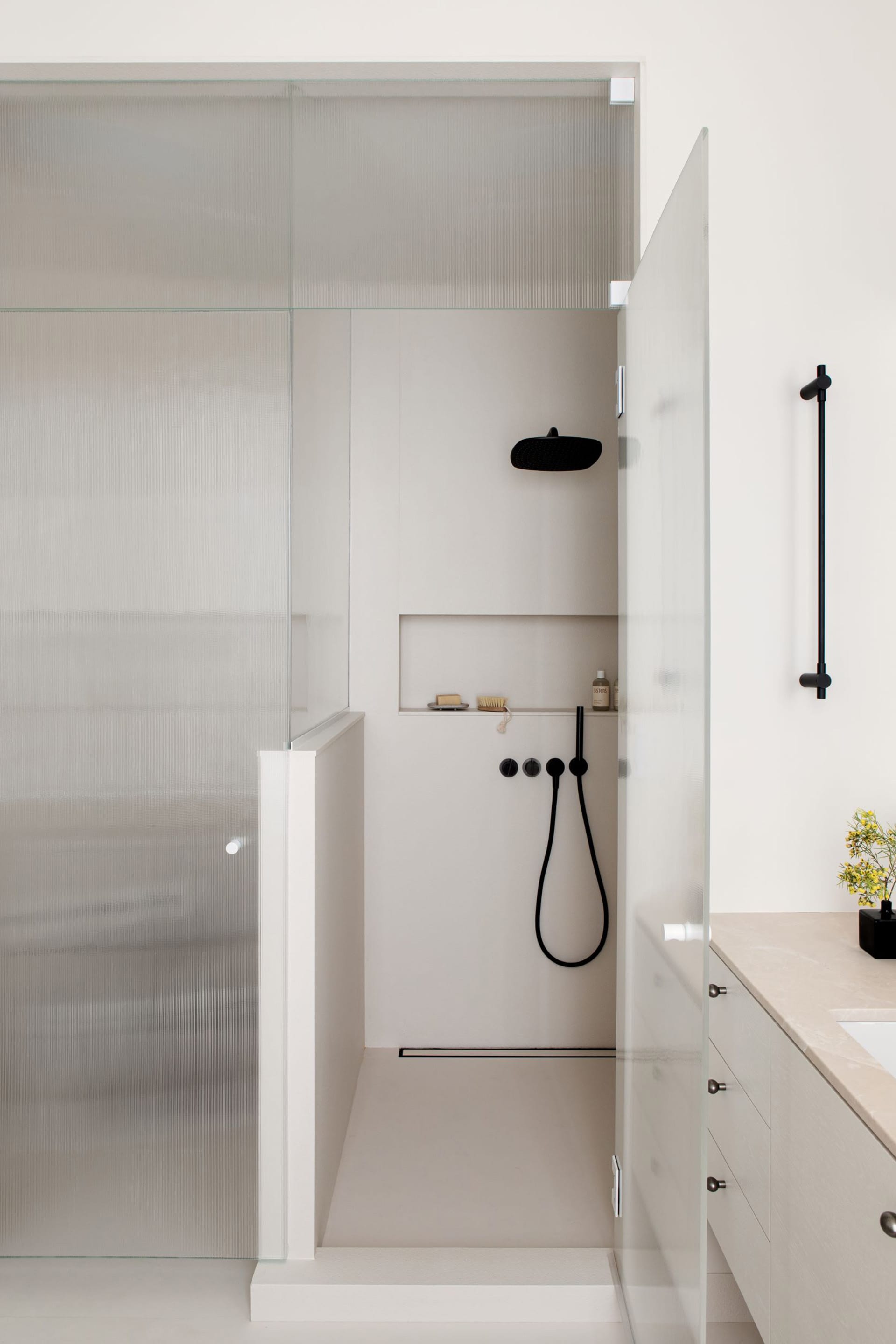 Shower in a primary bath with a glass door, black fixtures, cream counters, and white walls and floors.