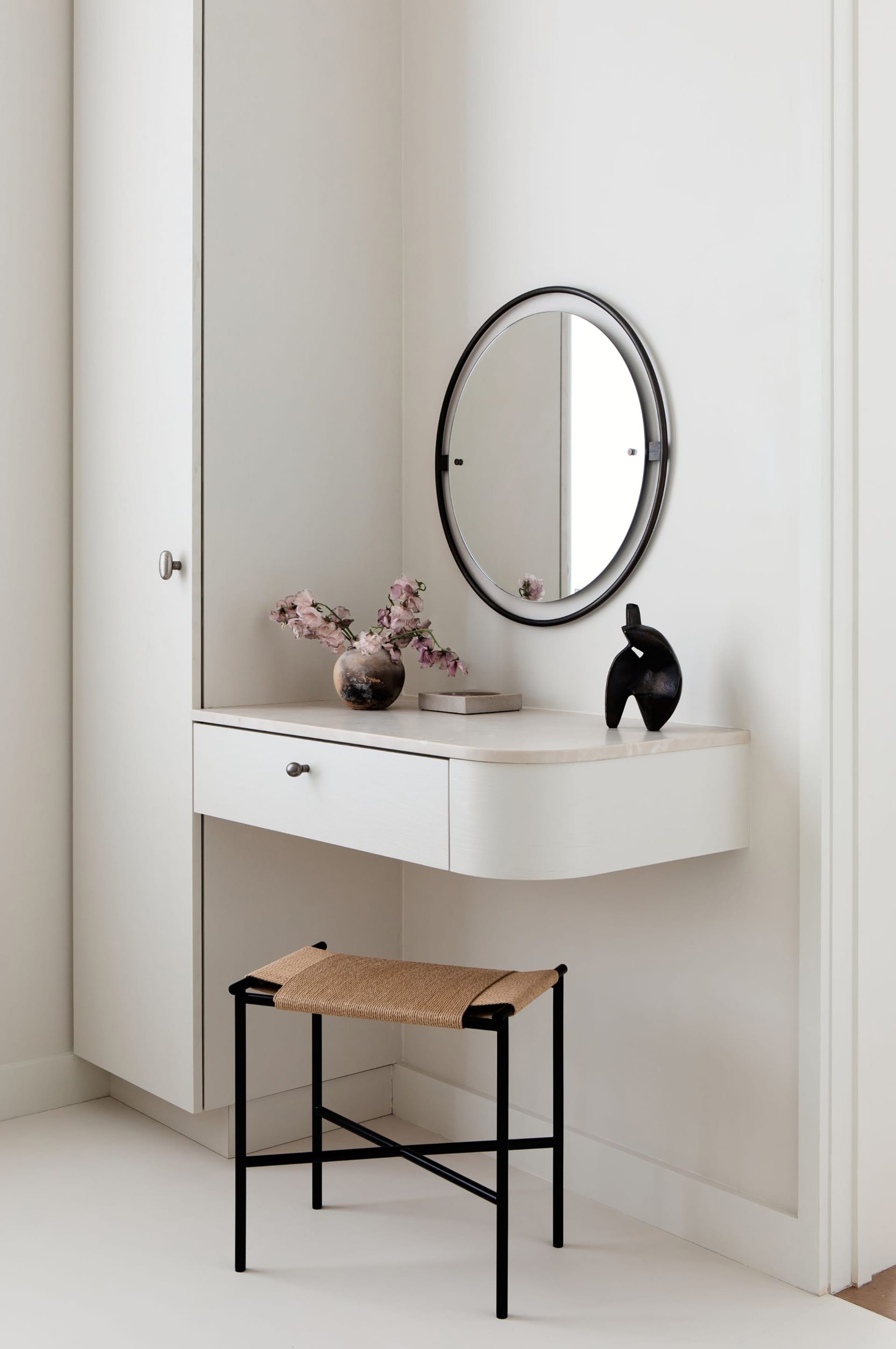 Vanity with a black mirror, statute, and wicker stool with a vase of flowers.