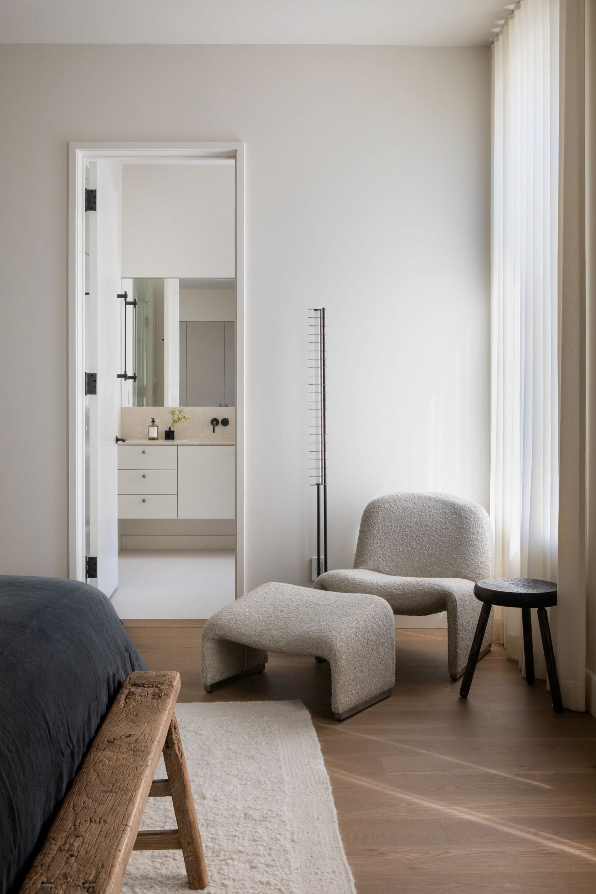 Bedroom with a wood bedframe and grey comforter. In the corner of the room, there's a matching chair and ottoman upholstered in light grey fabric. Through the open door, there is a primary bathroom with black fixtures, white walls and floors, and white cabinetry.