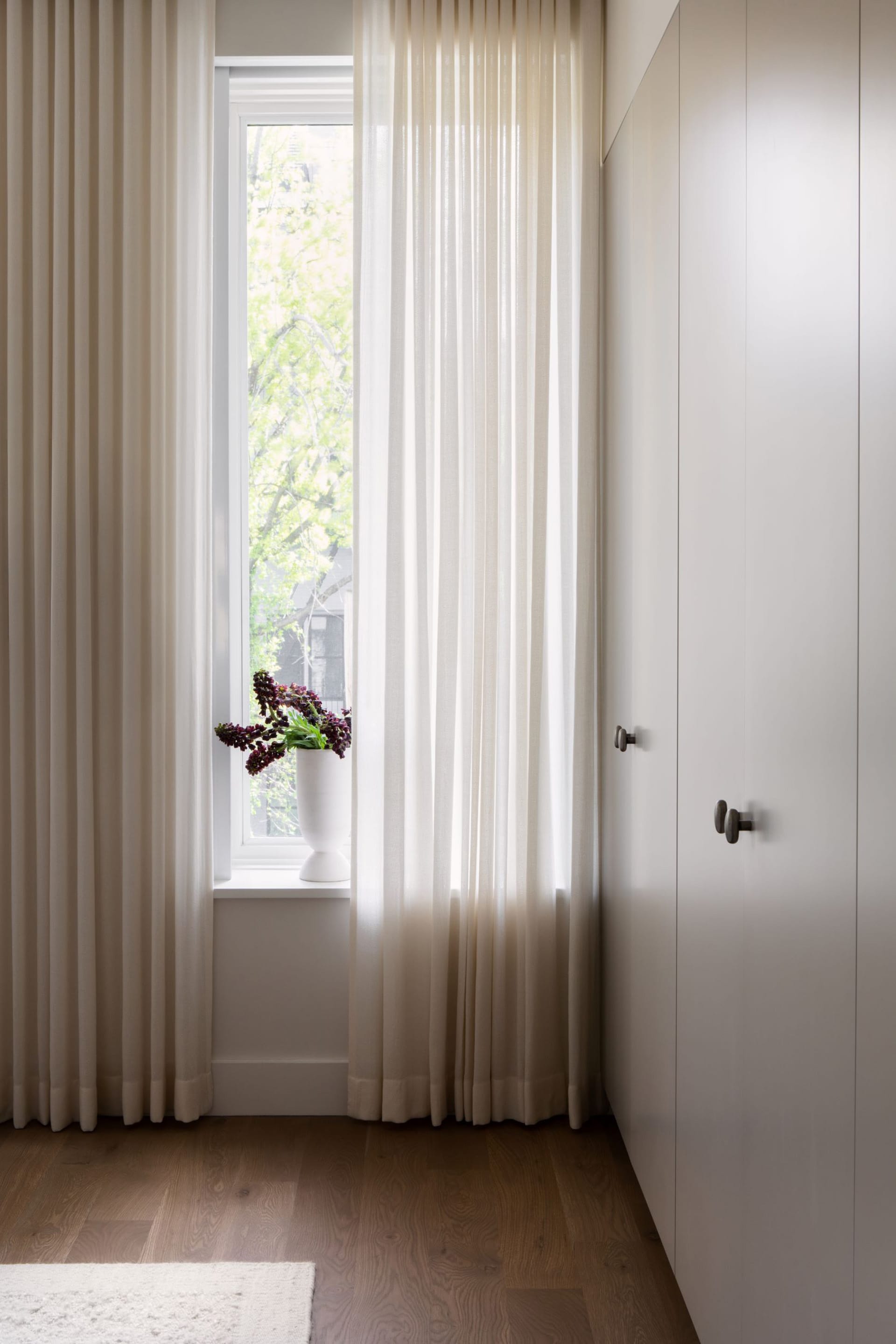 Custom closet doors next to a window with floor-to-ceiling curtains and a vase of flowers on the windowsill.